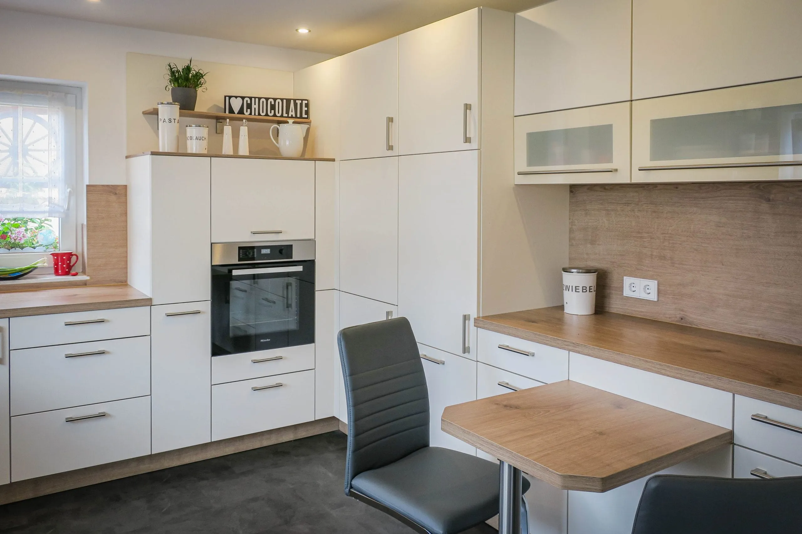 Weiße Küche mit Backofen-Hochschrank, Glas-Hängeschränken und Holz-Bartheke, an die ein kleiner Esstisch mit zwei grauen Lederstühlen anschließt