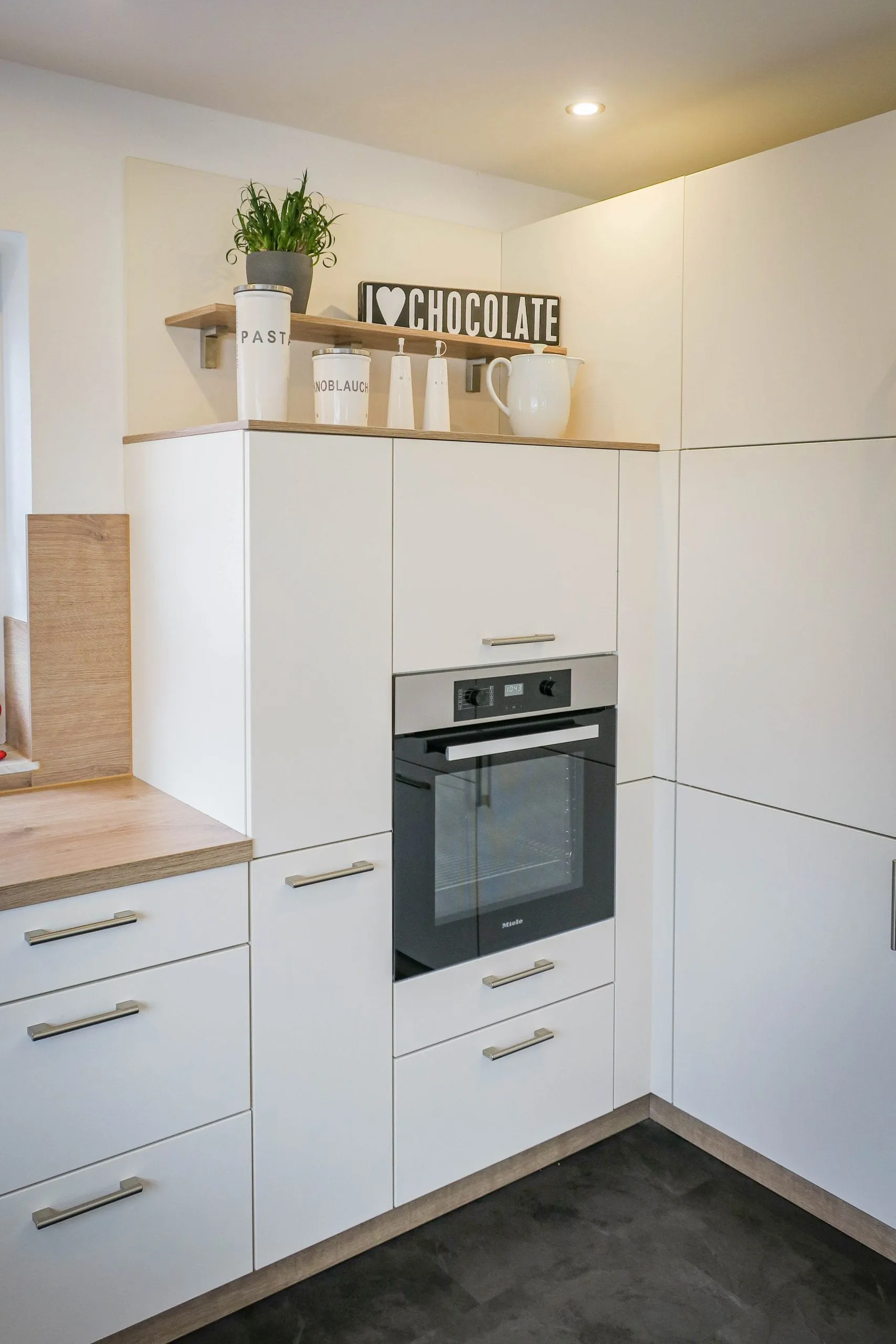 Weiße Eck-Küche mit Backofen-Hochschrank, Holz-Arbeitsplatte und Bartheke, kleinem Holz-Wandregal mit Vorratsdosen und dunklem Steinoptik-Boden