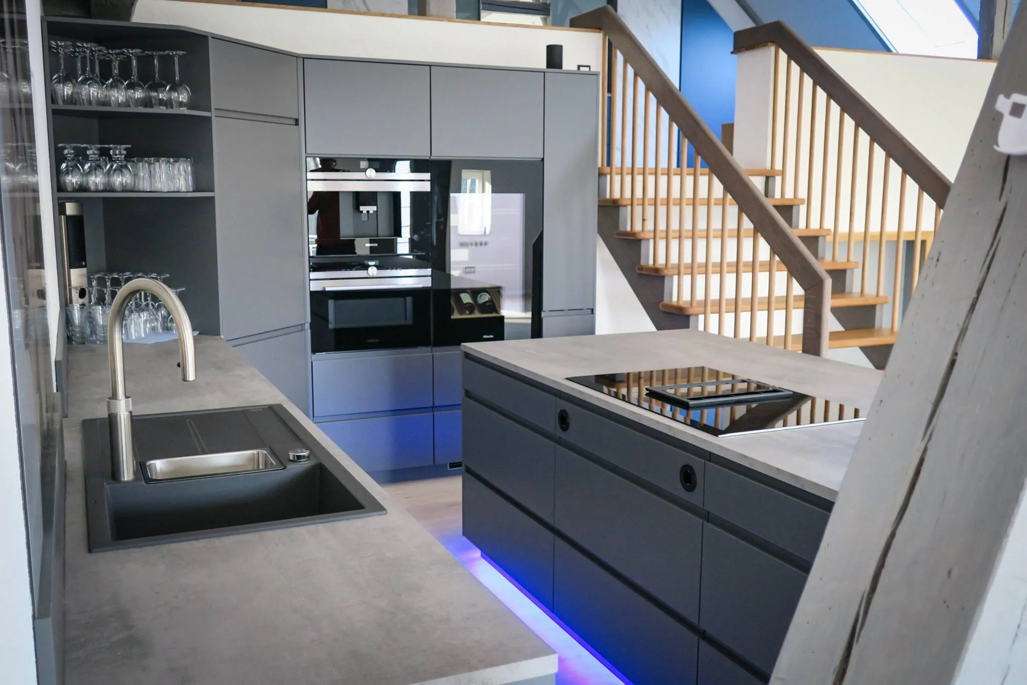 Anthrazitfarbene Küche mit Kochinsel mit Induktionskochfeld und blauer LED-Sockelbeleuchtung, Unterbau-Spüle mit Bogenarmatur, Hochschrankzeile mit Backofen und Glas-Hängeschrank neben einer Holztreppe