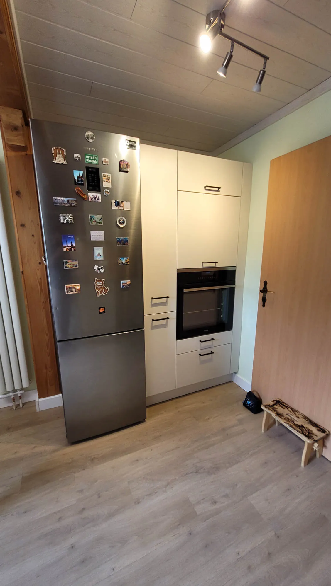 Edelstahl-Kühl-Gefrierkombination mit Magnetbild-Sammlung neben beigem Backofen-Hochschrank-Element, kleiner Holzbank an der Wand, helle Holzdecke