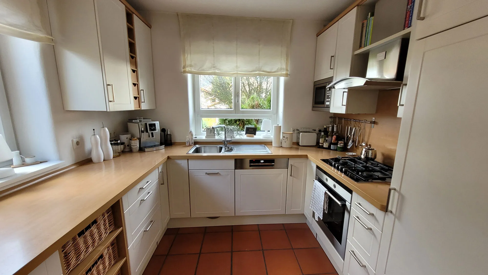 Vorher-Aufnahme der bestehenden Landhausküche mit magnolienfarbenen U-Fronten, Spüle am Fenster, Gas-Kochfeld und Backofen rechts, Holz-Arbeitsplatte und terrakottafarbener Fliesenboden