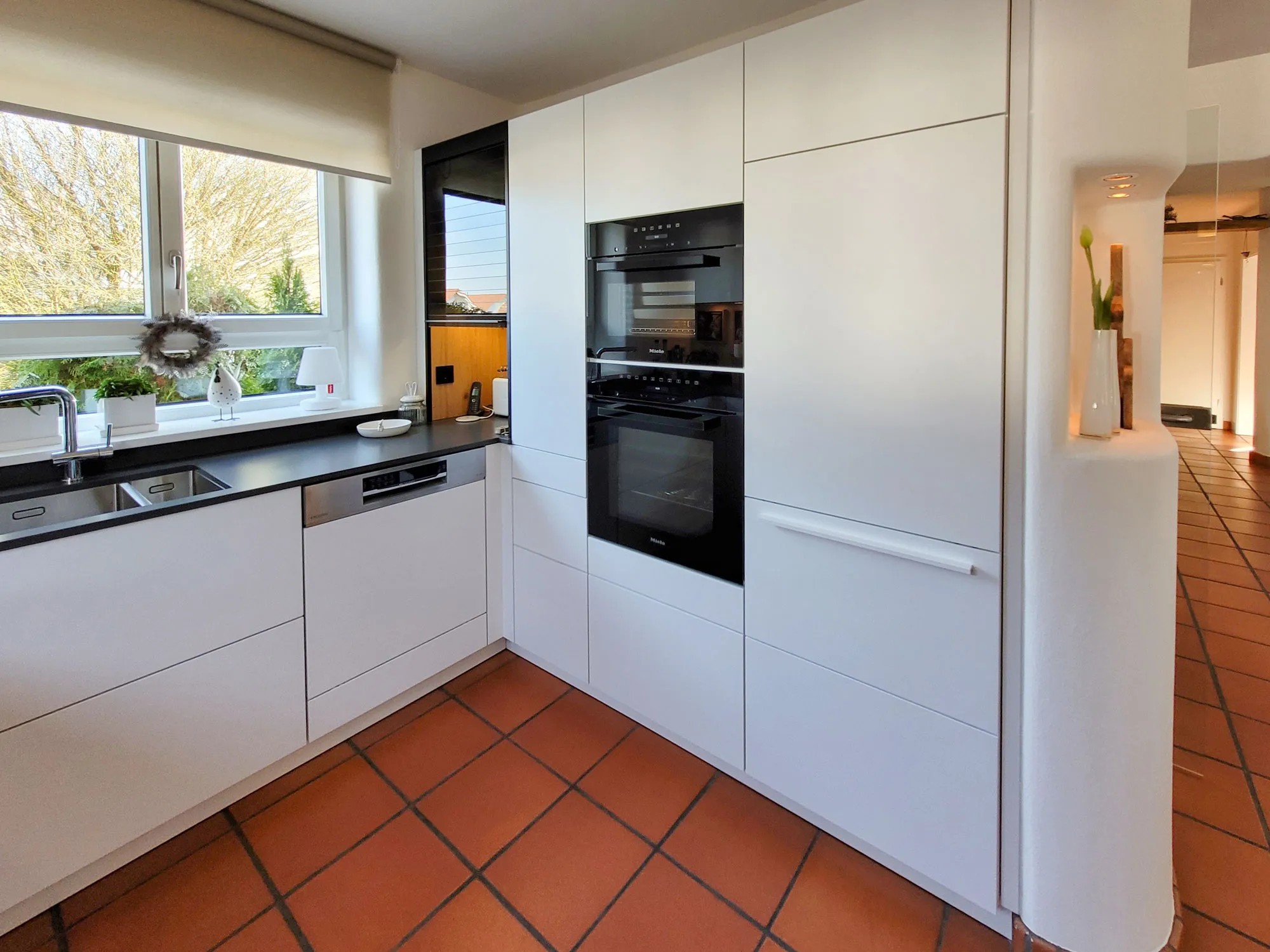 Weiße L-Küche mit grifflosen Fronten, anthrazitfarbener Arbeitsplatte, Spüle am Fenster und Backofen-Hochschrank-Kombination, terrakottafarbener Fliesenboden