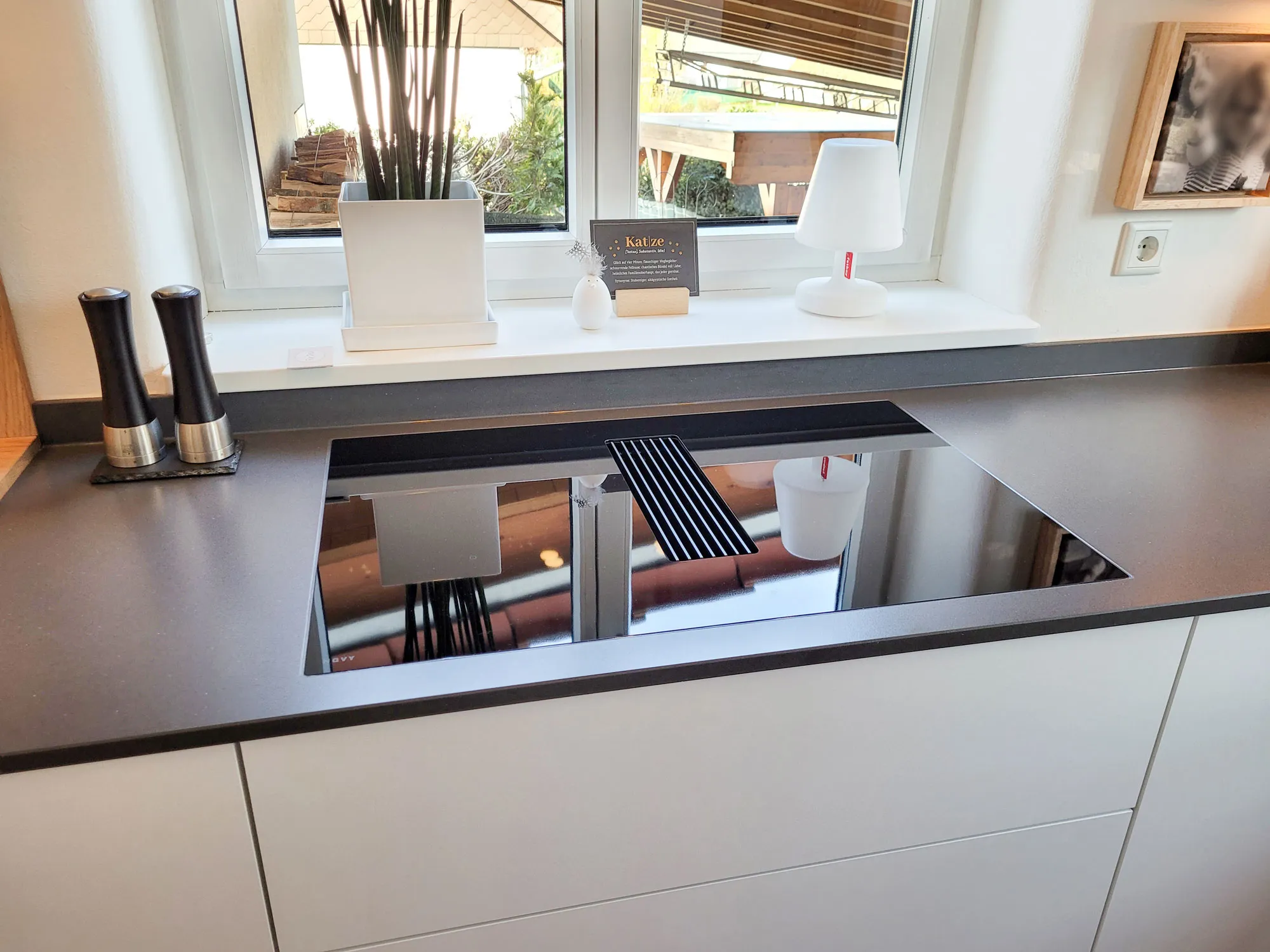 Detail Induktionskochfeld mit ausgefahrenem Muldenlüfter-Filter und sichtbarer Edelstahl-Konstruktion, anthrazitfarbene Arbeitsplatte vor dem Fensterbrett