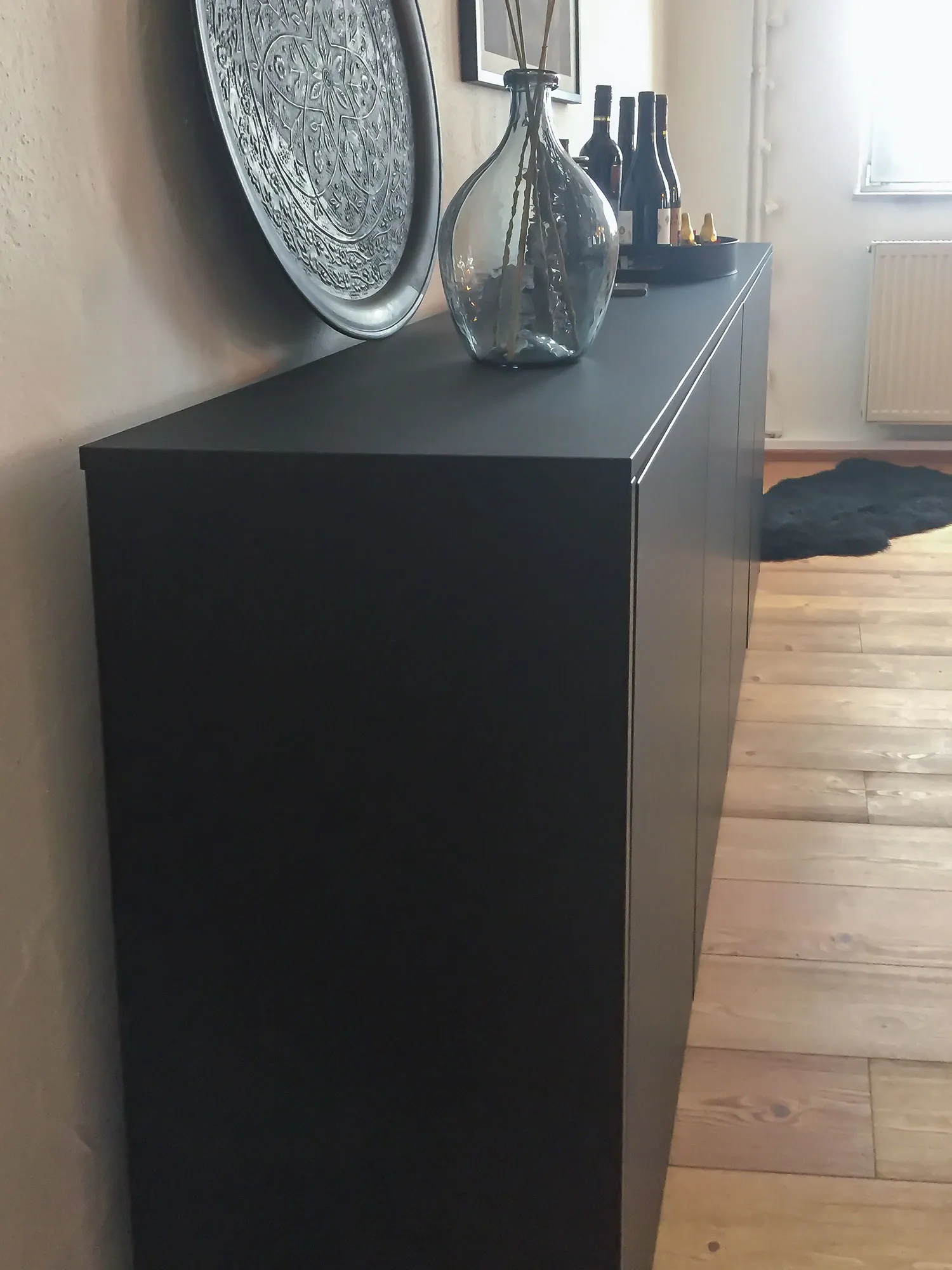 Detailaufnahme schwarzes Sideboard mit Glasvase, Ornament-Wandteller und Weinflaschen