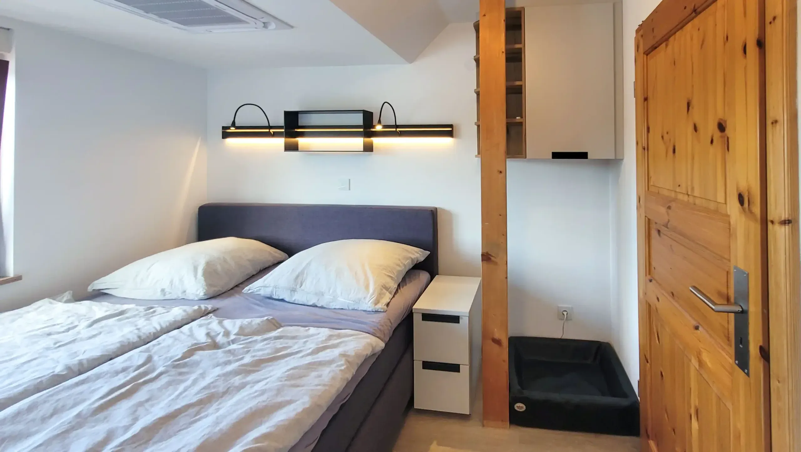 Schlafzimmer mit Doppelbett, schmalem Nachttisch, Holz-Regalwand und massiver Holztür.