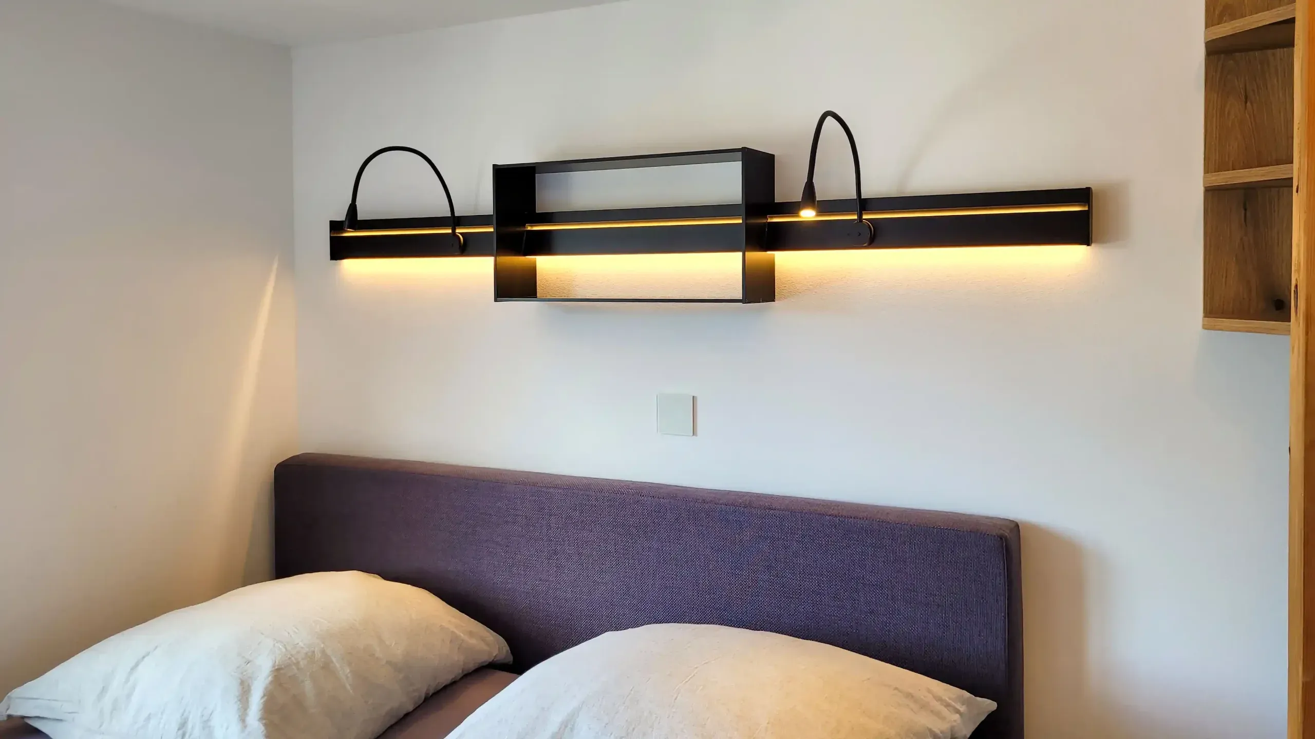 Wandleuchten-Element mit warmer LED-Beleuchtung über gepolstertem Bettkopfteil in dunklem Lila.