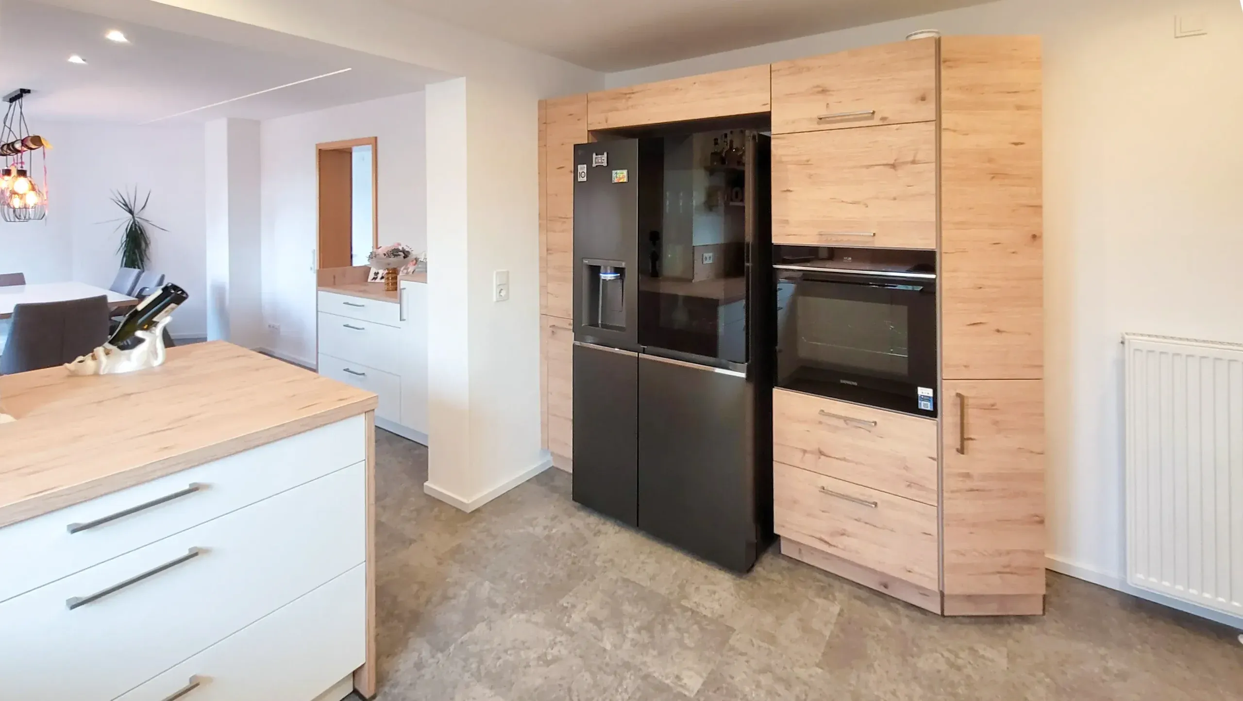 Holz-Hochschrankwand mit eingebautem schwarzem Side-by-Side-Kühlschrank, Backofen-Hochschrank und Dampfgarer, im Vordergrund die weiße Kochinsel mit Holz-Arbeitsplatte