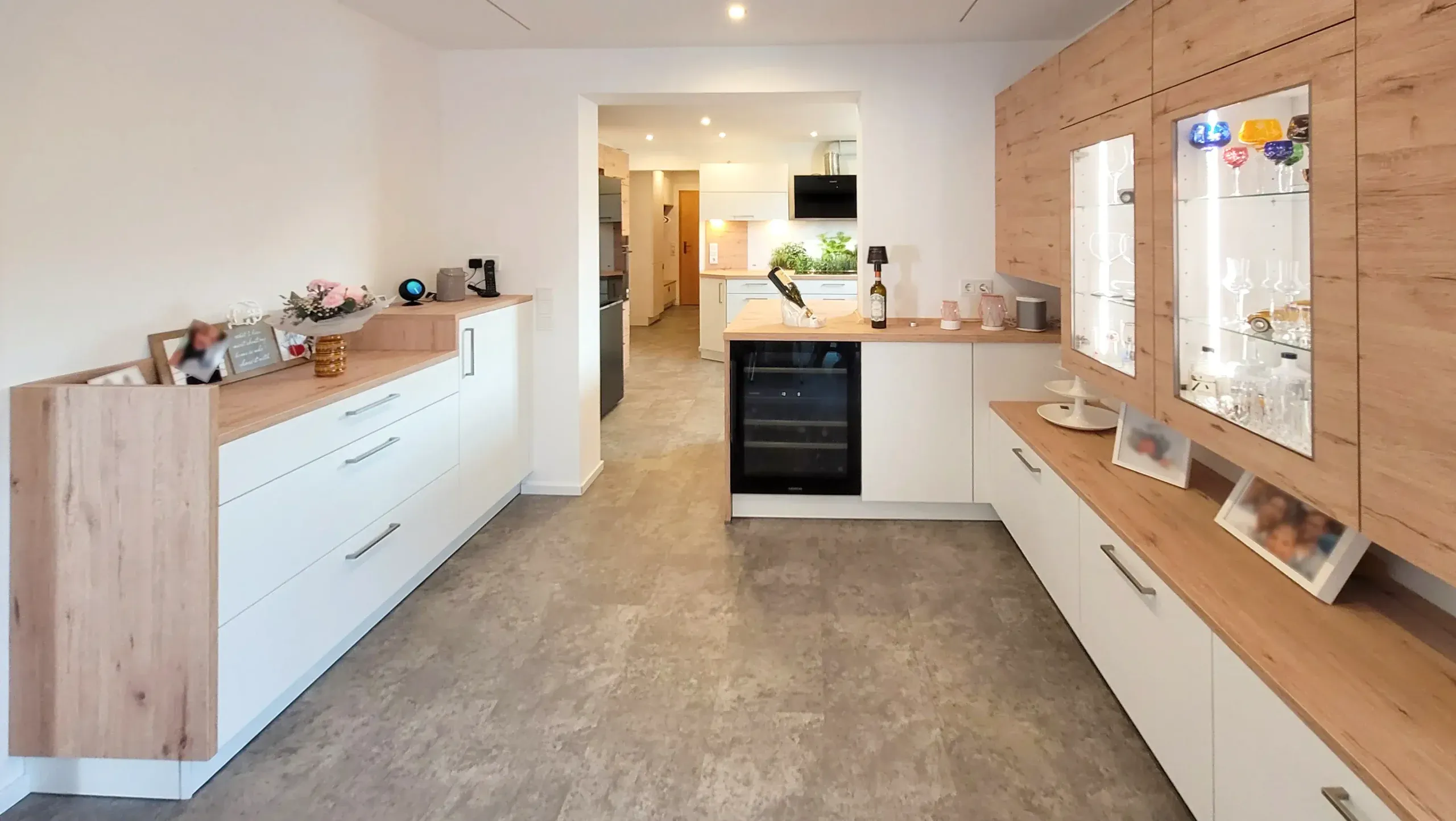 Wohnbereich mit zwei beleuchteten Glasvitrinen, Sideboard mit integriertem Weinkühlschrank, Bilderbord und Holz-Hochschrank, im Hintergrund Blick in die offene Küche