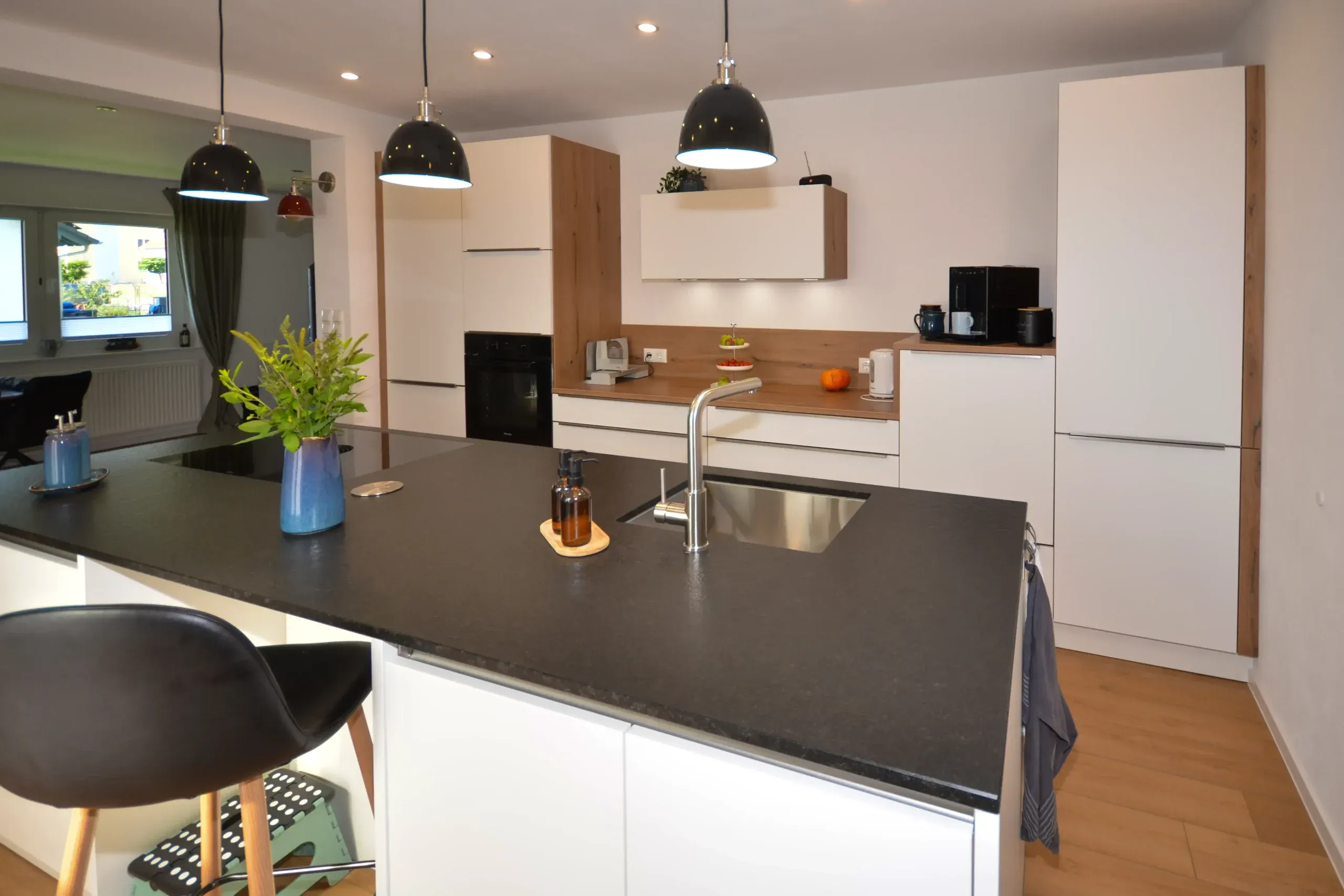 Moderne Wohnküche mit Kochinsel mit dunkler Arbeitsplatte, Spüle und Vase mit grünen Pflanzen, Wildeiche-Nischenrückwand, raumhohen weißen Schränken mit integrierten Backofen-Türmen, drei schwarzen Pendelleuchten und Barhocker im Vordergrund – Referenzprojekt von Broßler Küche Aktiv von Dietmar Kraus