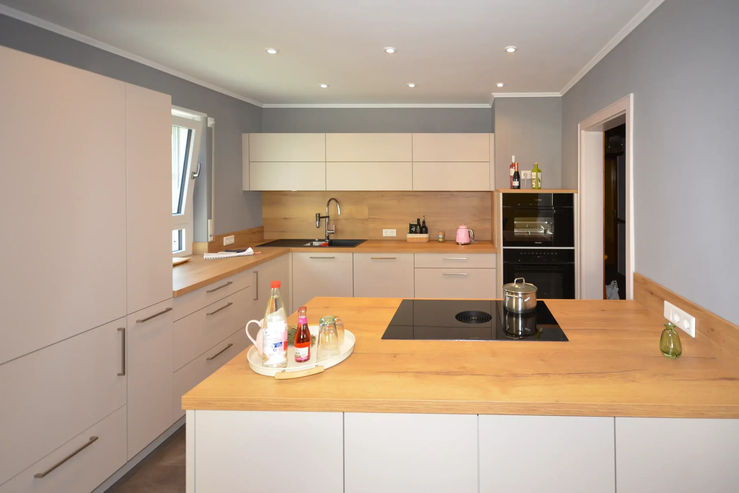 Moderne Küche mit Kochinsel im Vordergrund mit massiver Eichen-Arbeitsplatte und Kochfeld, dahinter Küchenzeile mit Spüle, beige/taupefarbenen matten Fronten, Holz-Nischenrückwand und integriertem Backofen – Referenzprojekt von Broßler Küche Aktiv von Dietmar Kraus