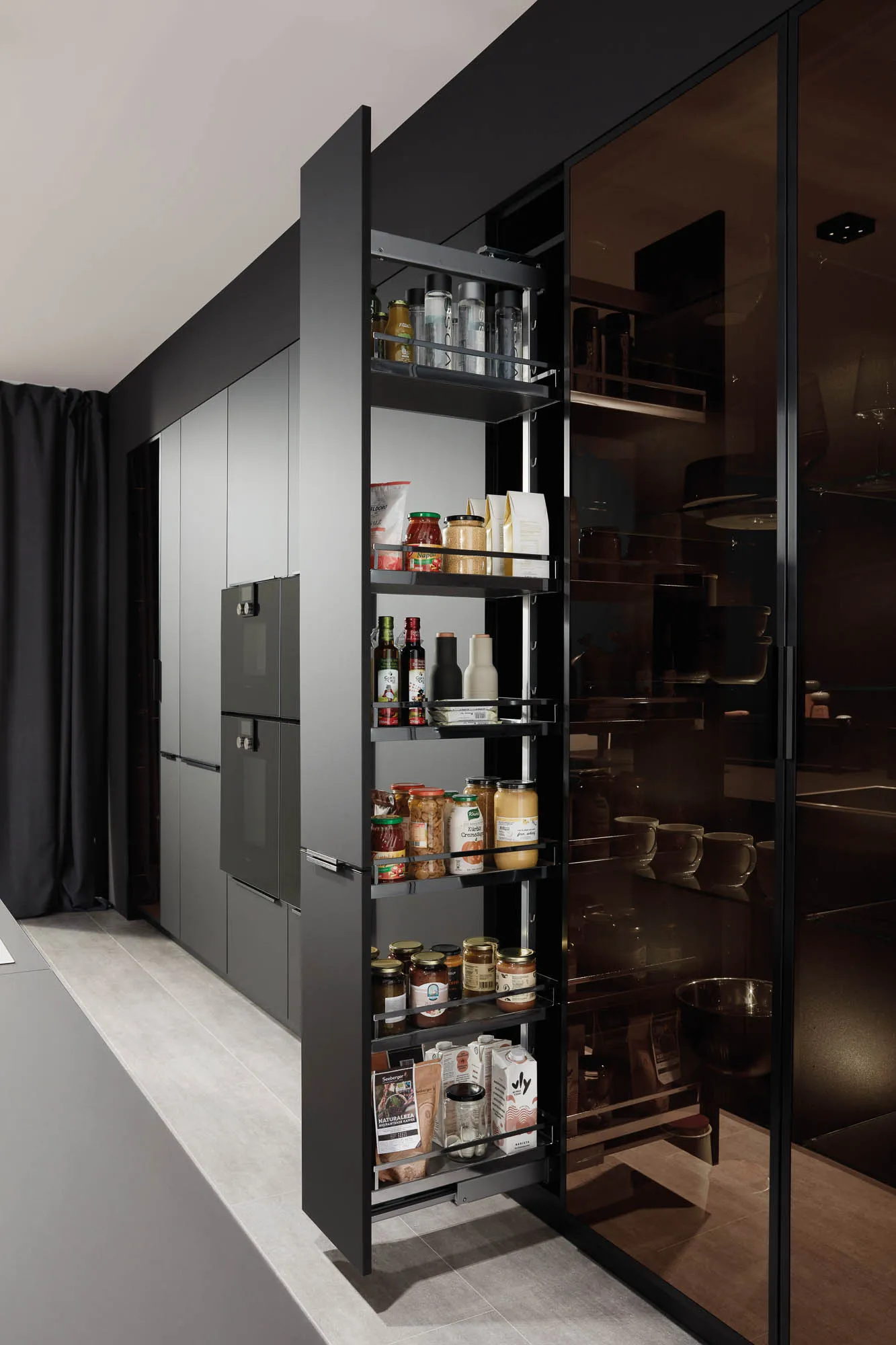 Küchenrückwand in mattem Grau mit geöffnetem Apothekerschrank, rechts Glastüren, im Hintergrund schwarze Backöfen.