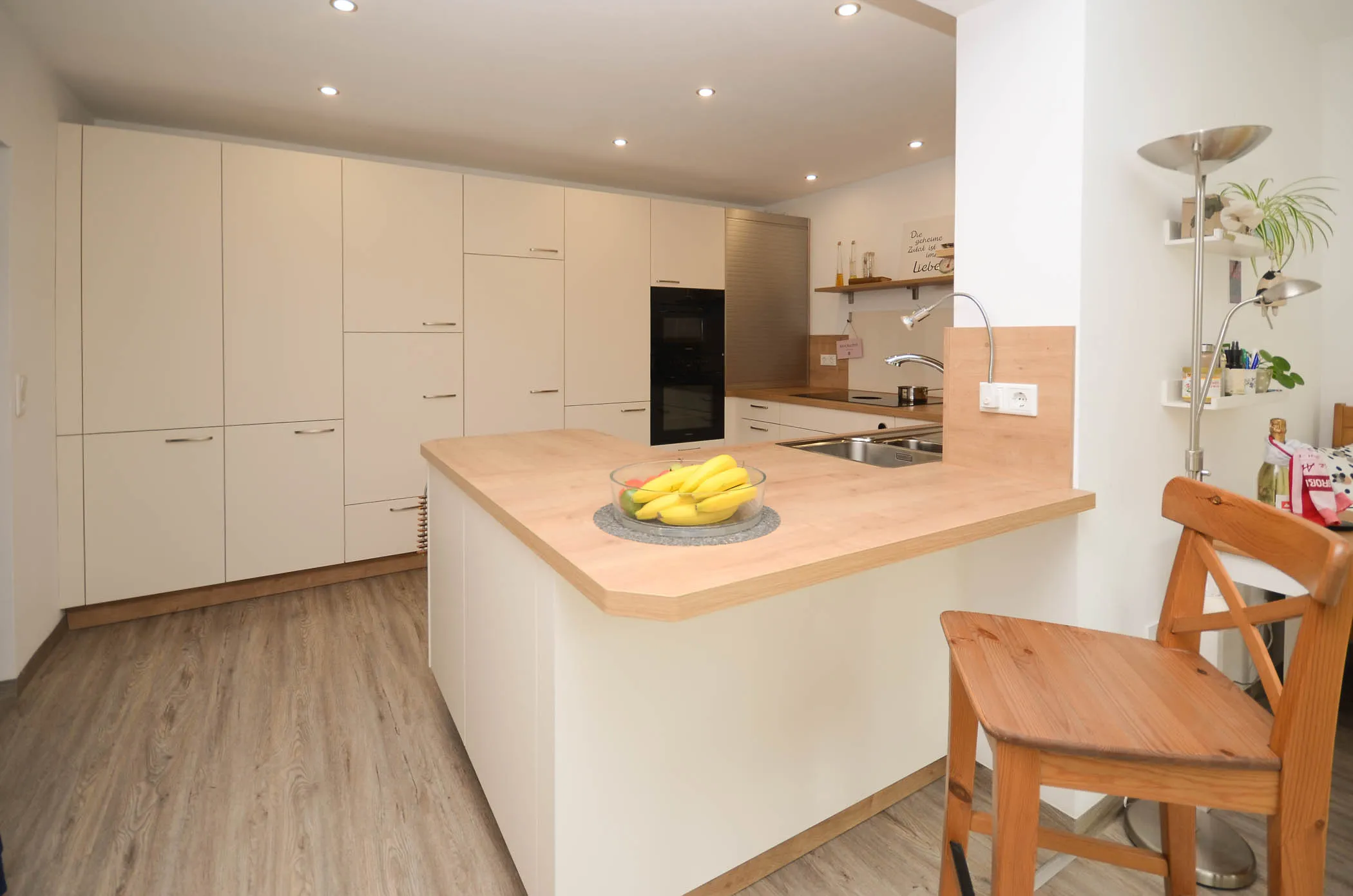 Helle L-Küche mit weißen Hochglanzfronten, Halbinsel mit heller Holz-Arbeitsplatte, integrierten Backofen-Hochschränken und Punktstrahlern an der Decke – Referenzprojekt von Broßler Küche Aktiv