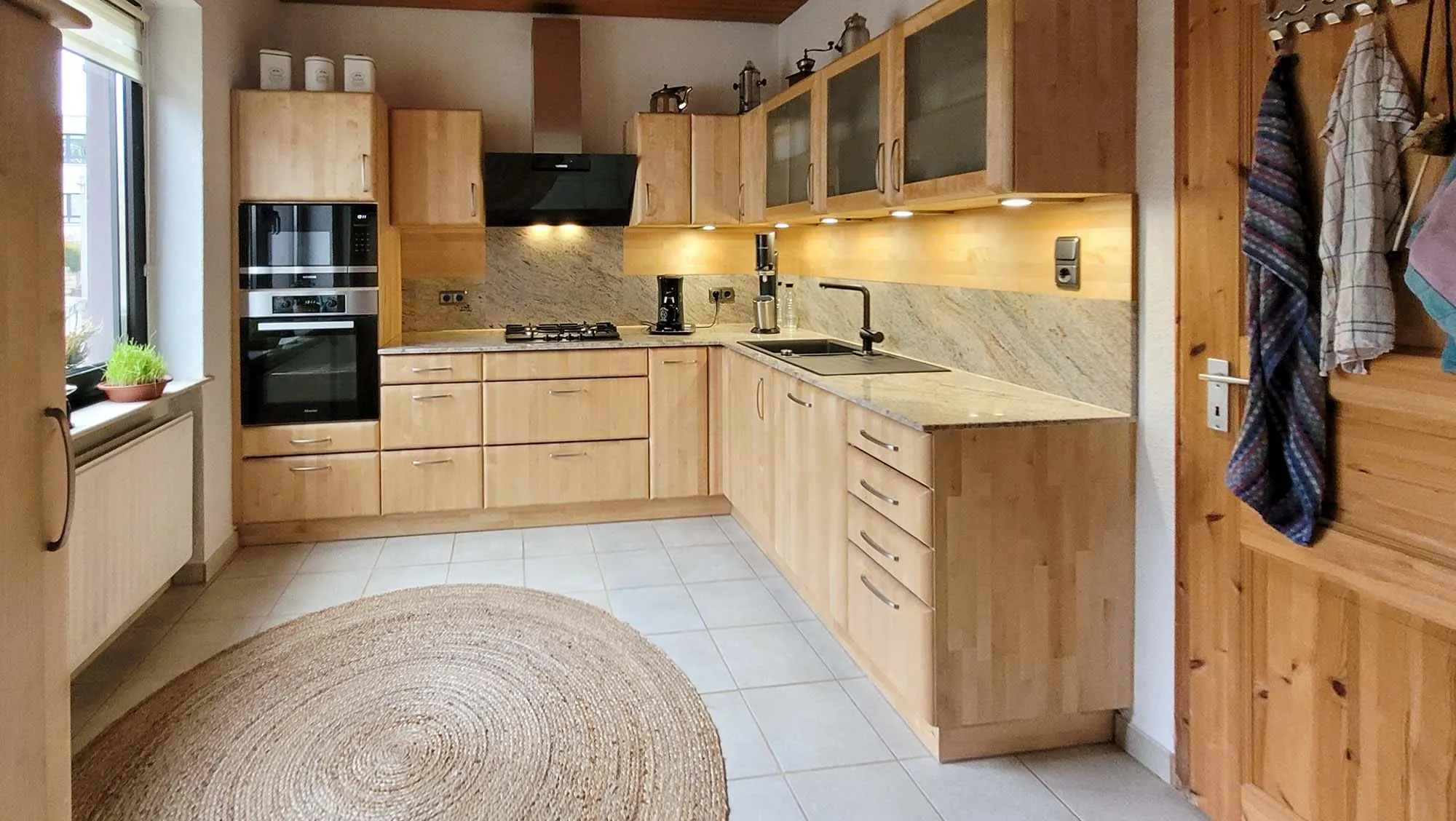 Gesamtansicht der hellen Eiche-L-Küche mit Backofen-Hochschrank, schwarzem Wand-Dunstabzug, Gaskochfeld, Spüle im Eckbereich, Glas-Hängeschränken und Sisalteppich am Boden