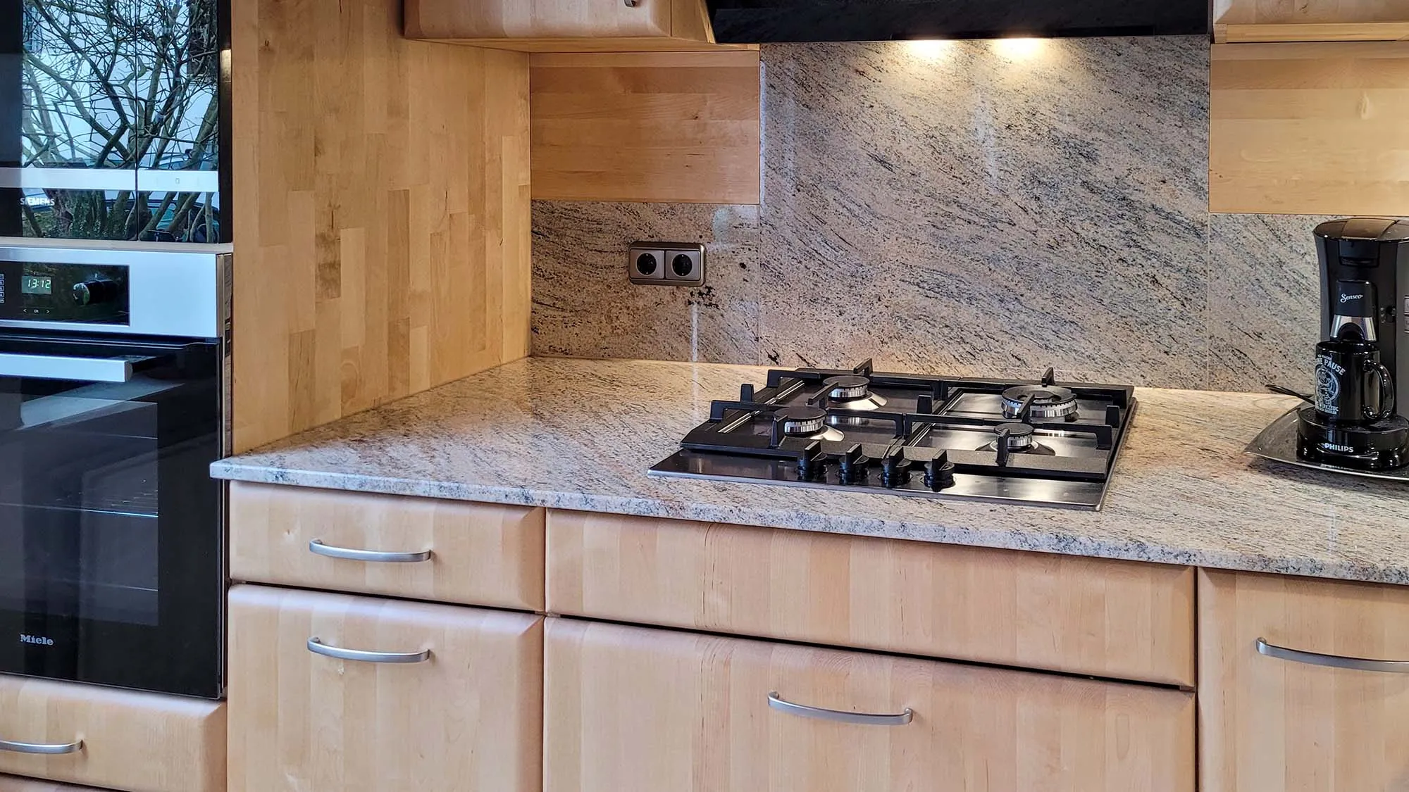 Detail Kochbereich mit schwarzem 5-Flammen-Gaskochfeld auf Granit-Arbeitsplatte, integrierten Steckdosen in der Rückwand, Backofen-Hochschrank links und Kaffeevollautomat rechts