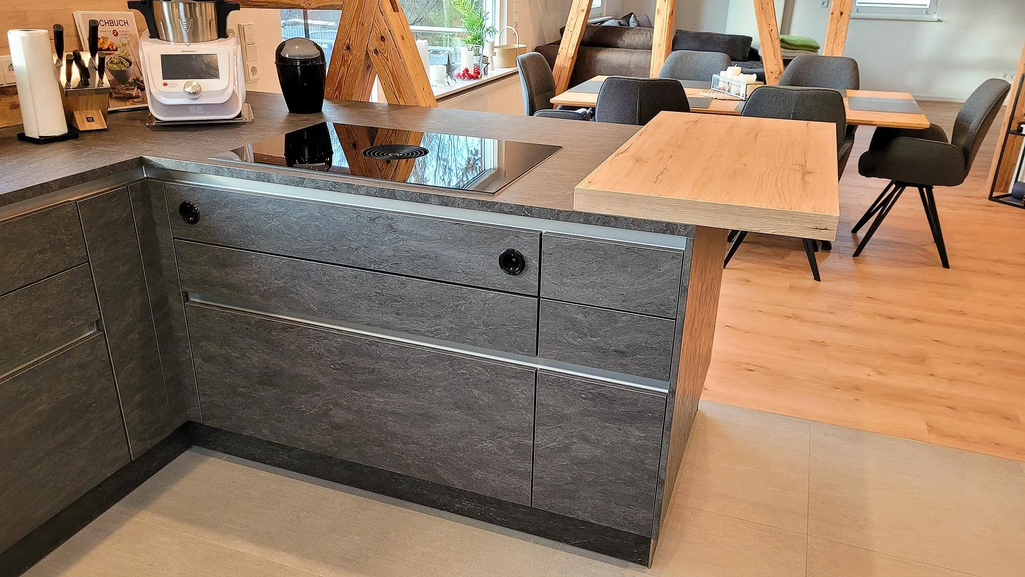 Dunkelbraune Kochinsel mit angesetzter Holz-Bartheke und Induktionskochfeld, im Hintergrund Essbereich mit langem Esstisch und schwarzen Polsterstühlen
