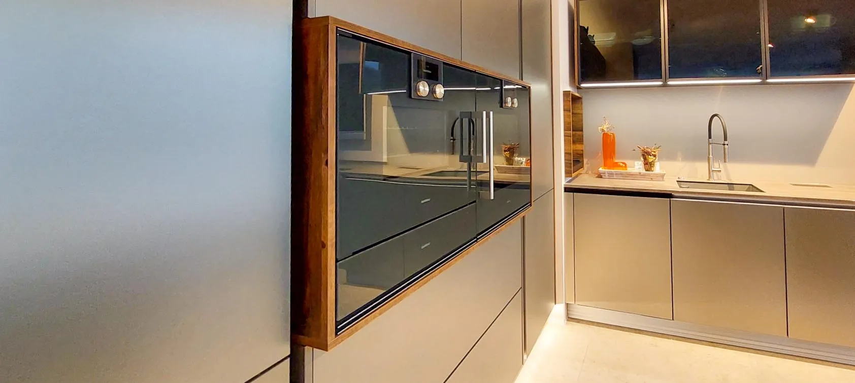 Frontalansicht einer Küchenzeile bei Abendlicht: grifflose anthrazitgraue Fronten, Holz-eingerahmter Backofen-Hochschrank und Spülbereich mit Bogenarmatur