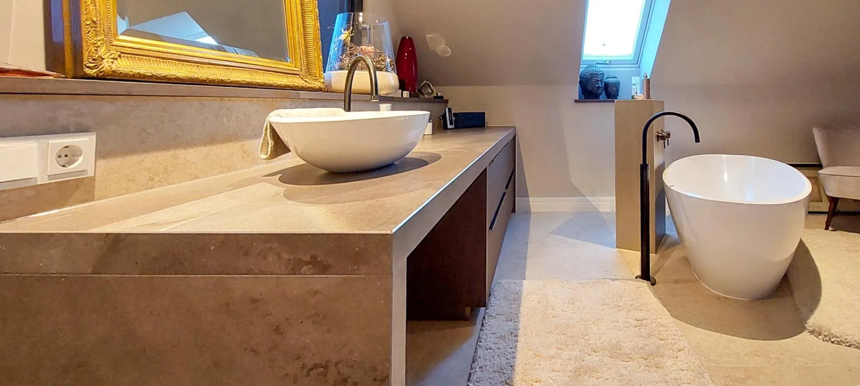 Badezimmer mit Naturstein-Waschtisch, weissem Aufsatzwaschbecken, vergoldetem Rahmenspiegel und freistehender Badewanne mit schwarzer Standarmatur