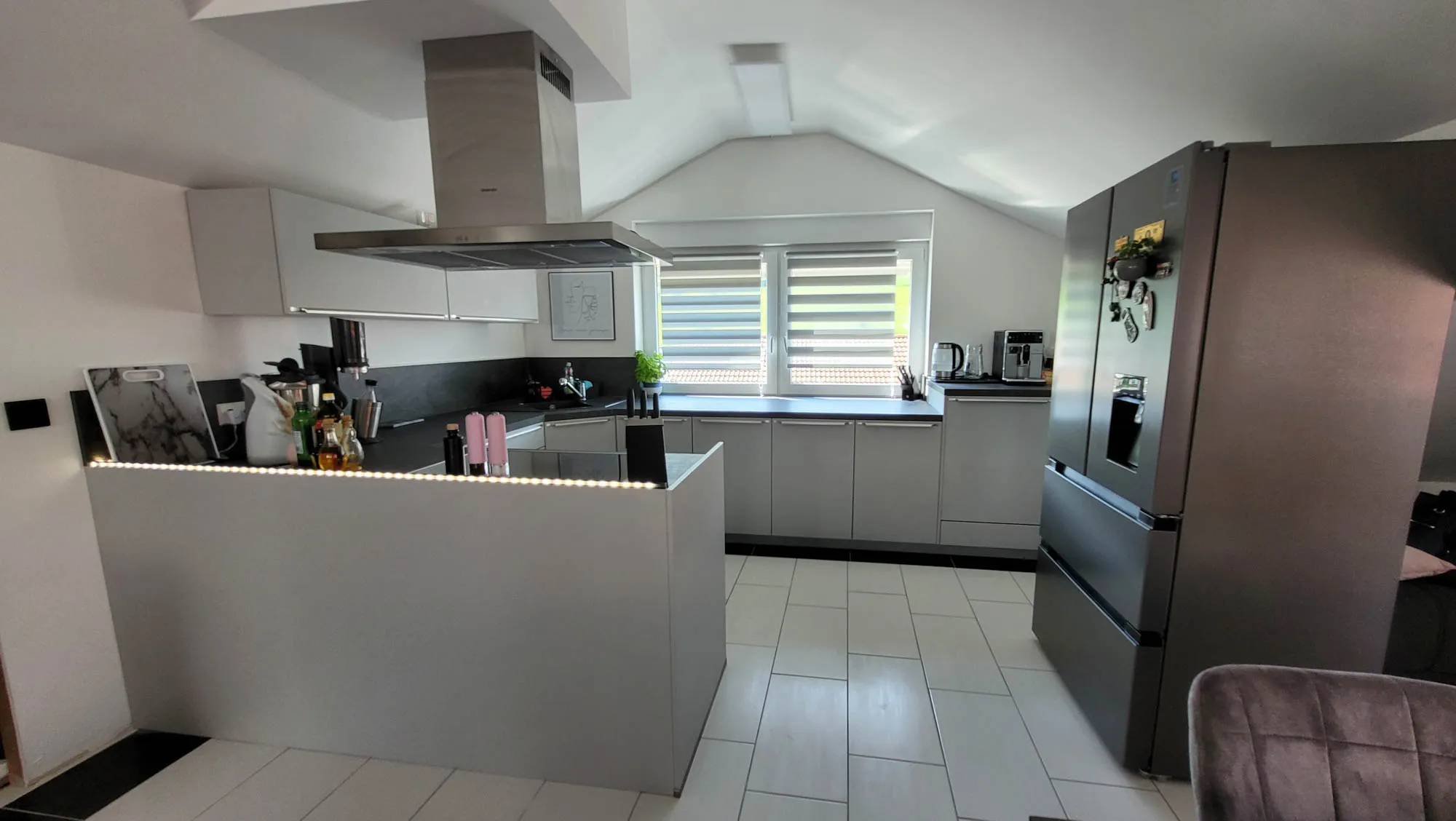 Helle Küche mit Halbinsel im Vordergrund (LED-Sockelbeleuchtung), Insel-Dunstabzug über dem Kochbereich, Arbeitszeile am Fenster und Side-by-Side-Edelstahlkühlschrank rechts
