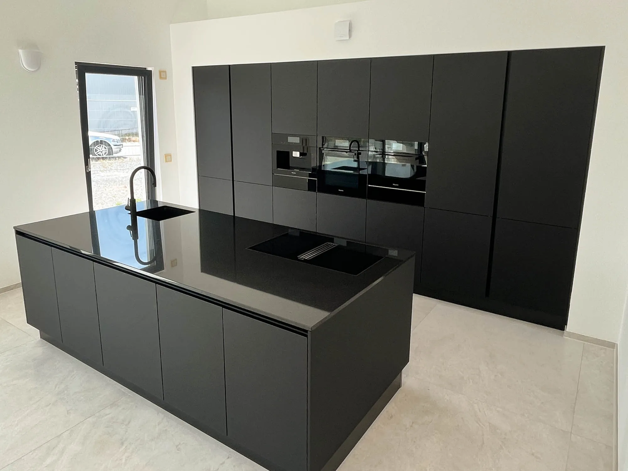 Schwarze grifflose Küche mit großer Kochinsel, schwarzer Steinarbeitsplatte und integriertem Induktionskochfeld mit Muldenlüfter, eingelassene Edelstahl-Spüle mit hoher schwarzer Armatur, im Hintergrund Hochschrankzeile mit Backofen und Mikrowelle, große Fenstertür nach draußen, heller Steinfliesenboden – Küchenplanung von Dietmar Kraus für Broßler Küche Aktiv