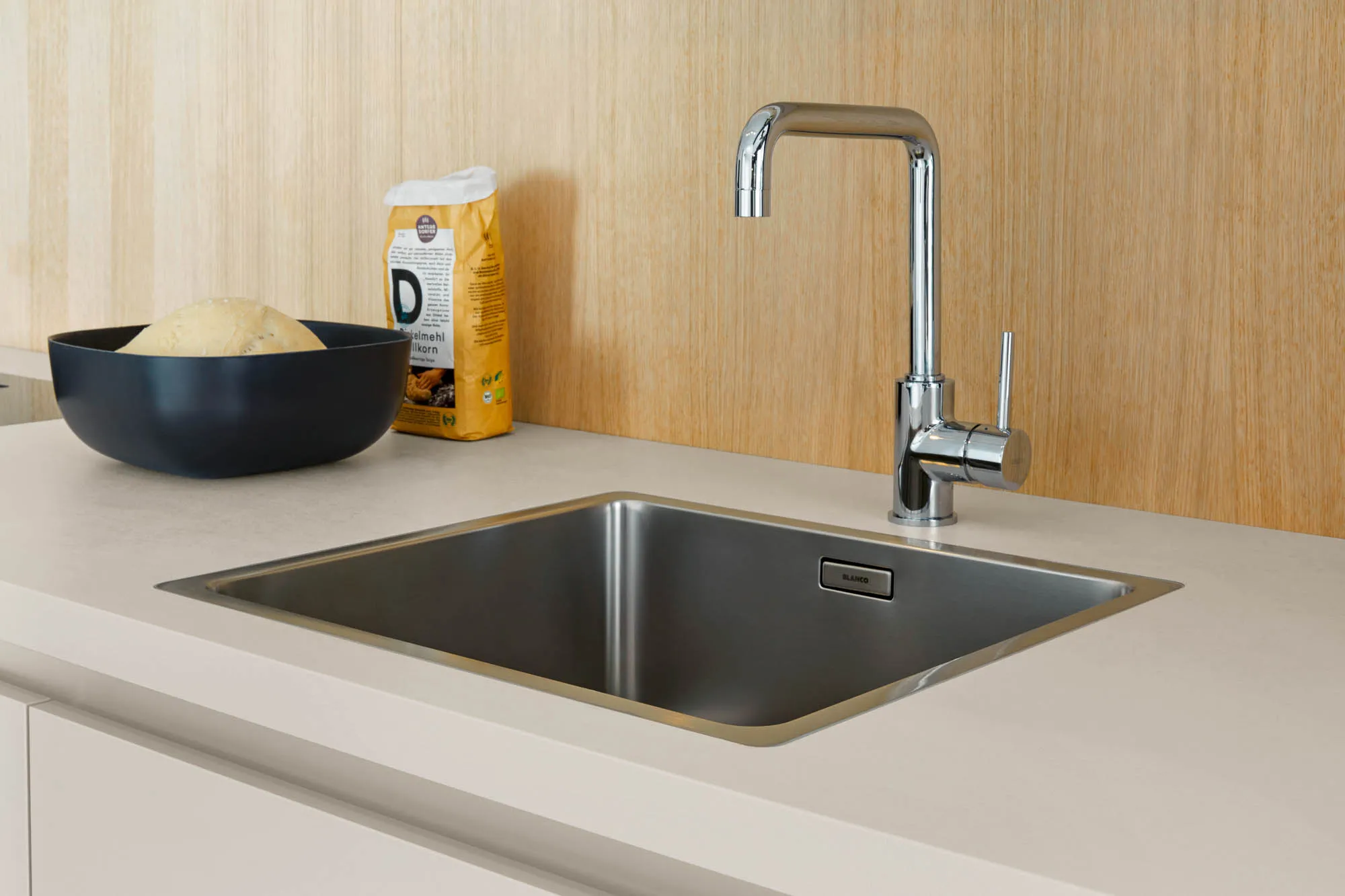 Flächenbündig eingebaute Edelstahlspüle mit Chromarmatur in der muschelweiß matten Arbeitsplatte einer grifflosen Küche vor Holzwand