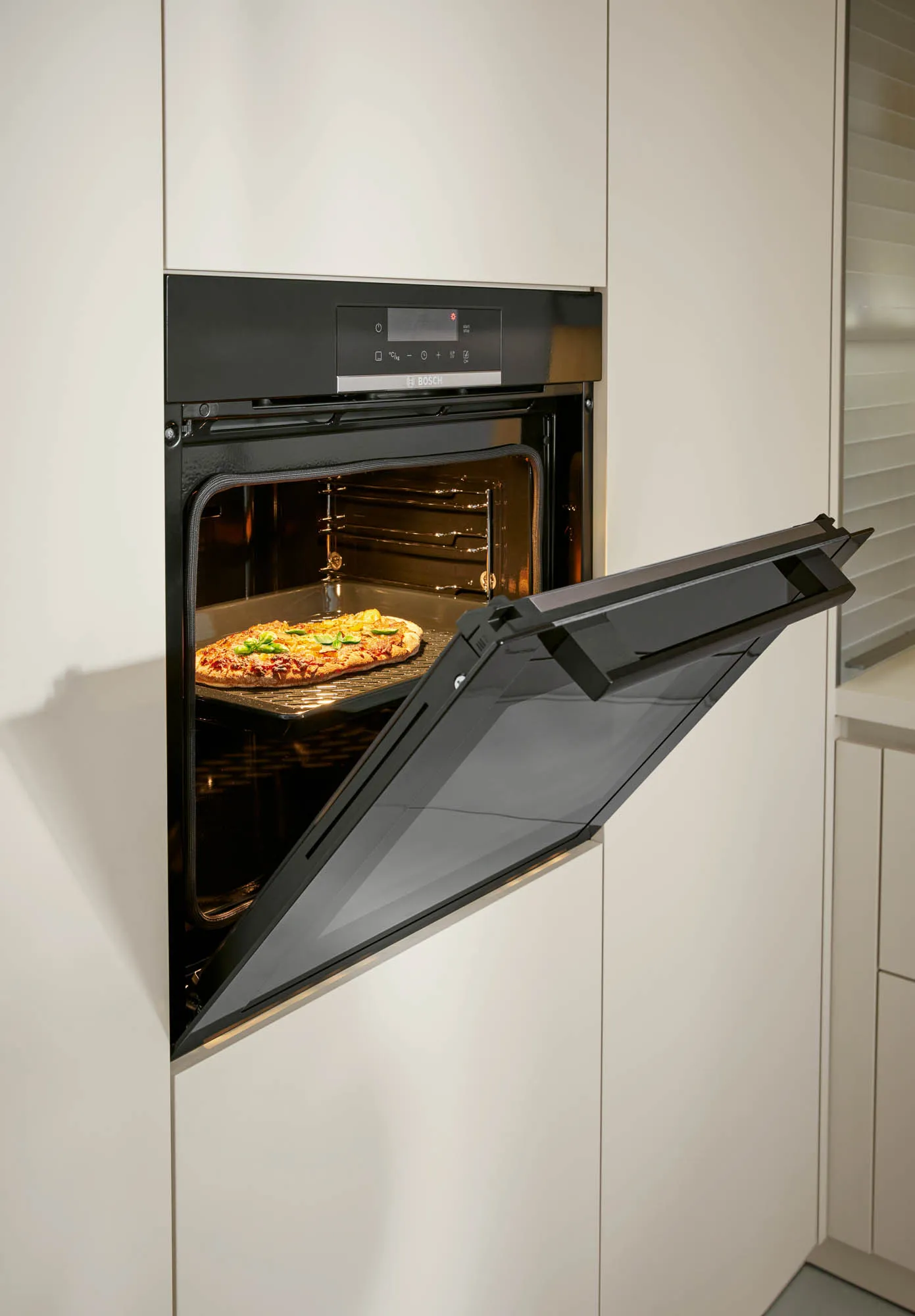 Geöffneter schwarzer Einbaubackofen in einem muschelweiß matten Hochschrank mit frisch gebackener Pizza auf dem Backblech