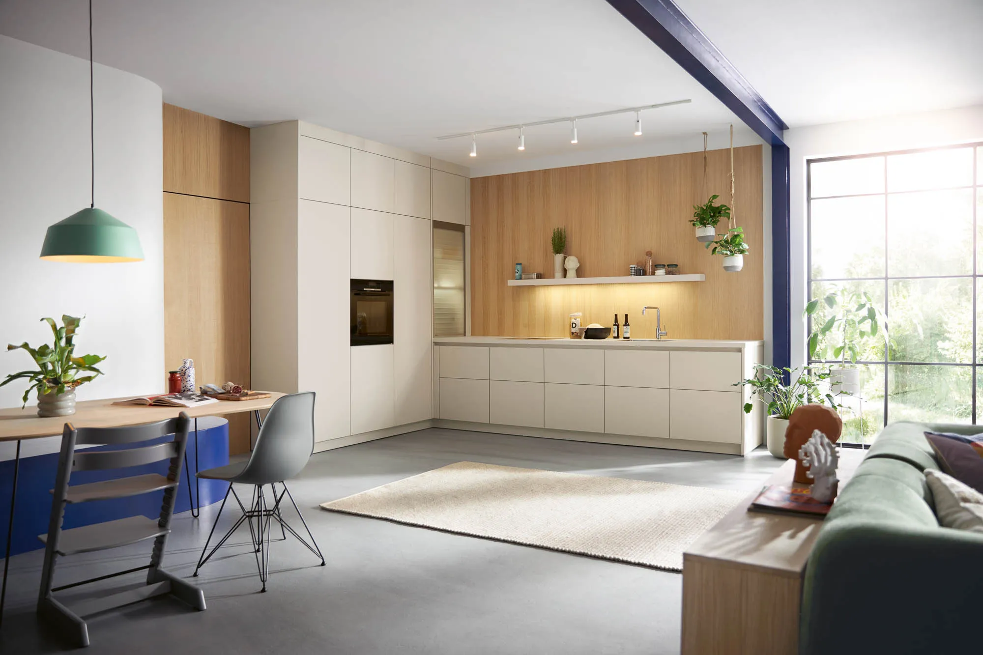 Offene Wohnküche mit griffloser L-Küche in Muschelweiß matt, deckenhoher Schrankzeile mit Backofen, Holzwand mit beleuchtetem Regal und Essbereich mit Holztisch