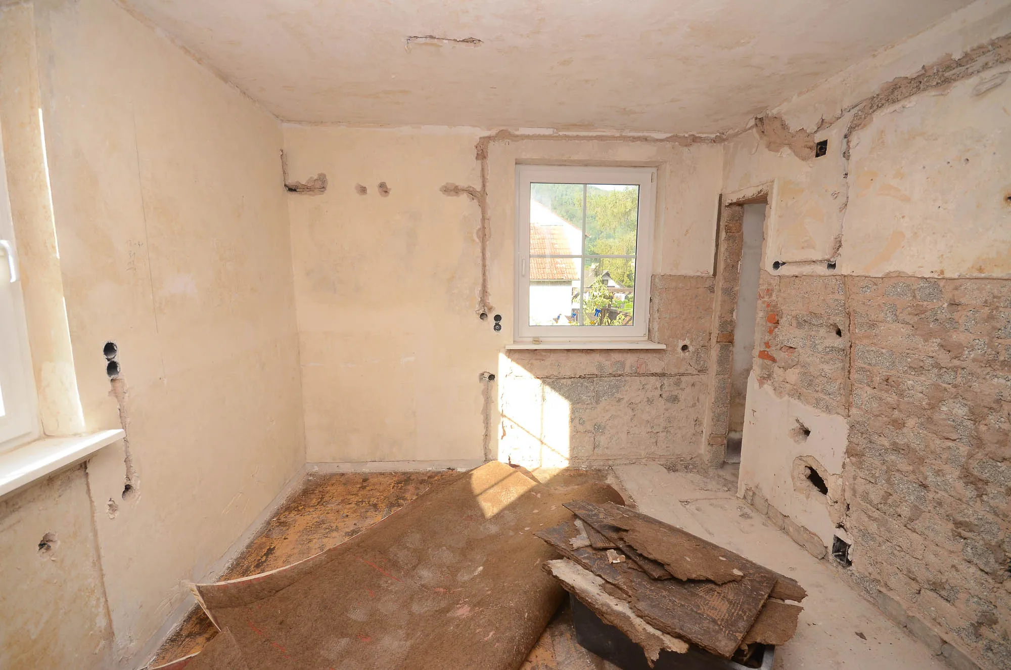 Küchenraum im Rohzustand vor der Renovierung mit freigelegten Wänden und entferntem Bodenbelag, Tageslicht durch ein Fenster