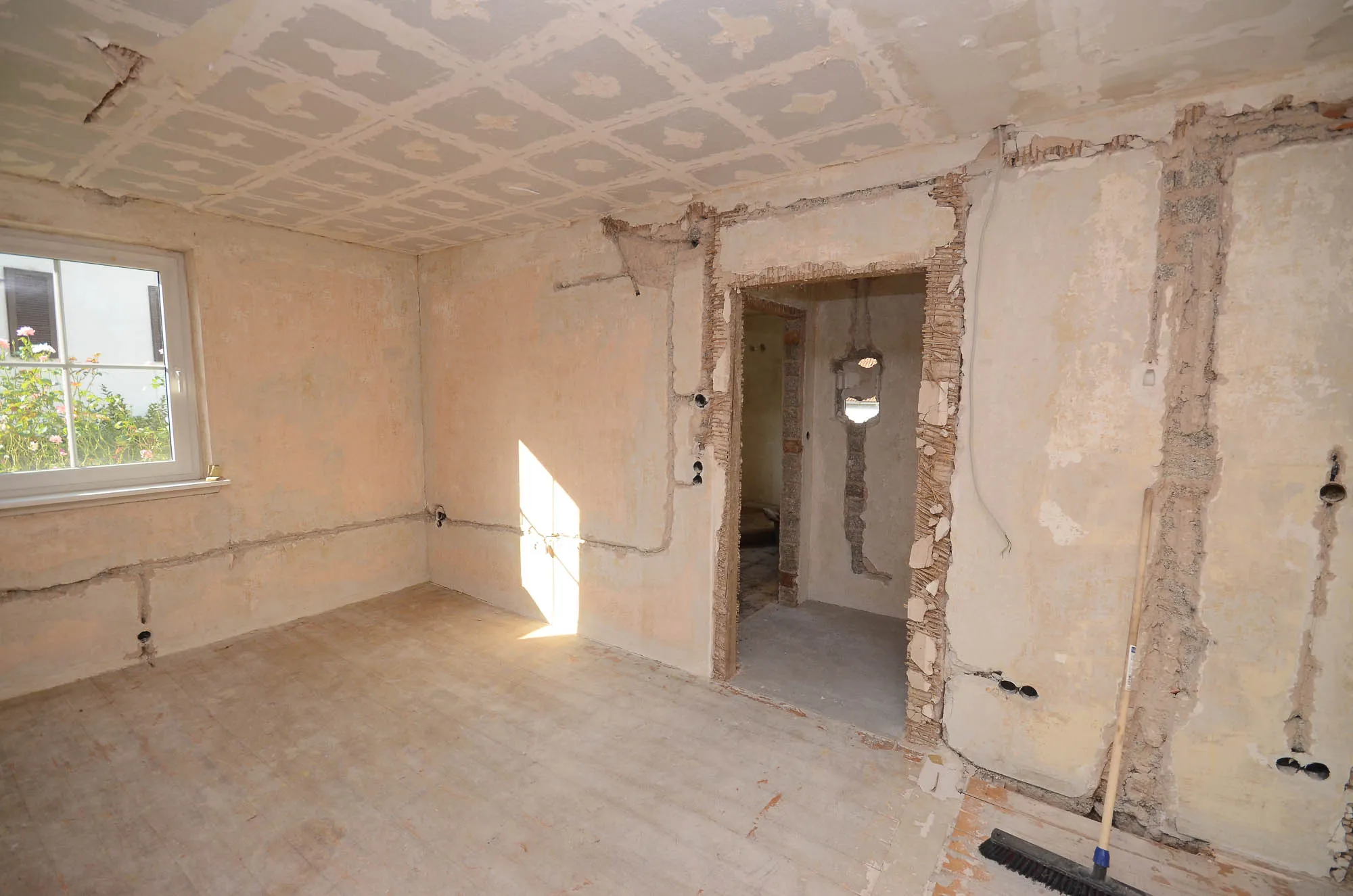 Raum im Rohbauzustand mit Wanddurchbruch, alter Deckenverkleidung und Tageslicht durch ein Fenster