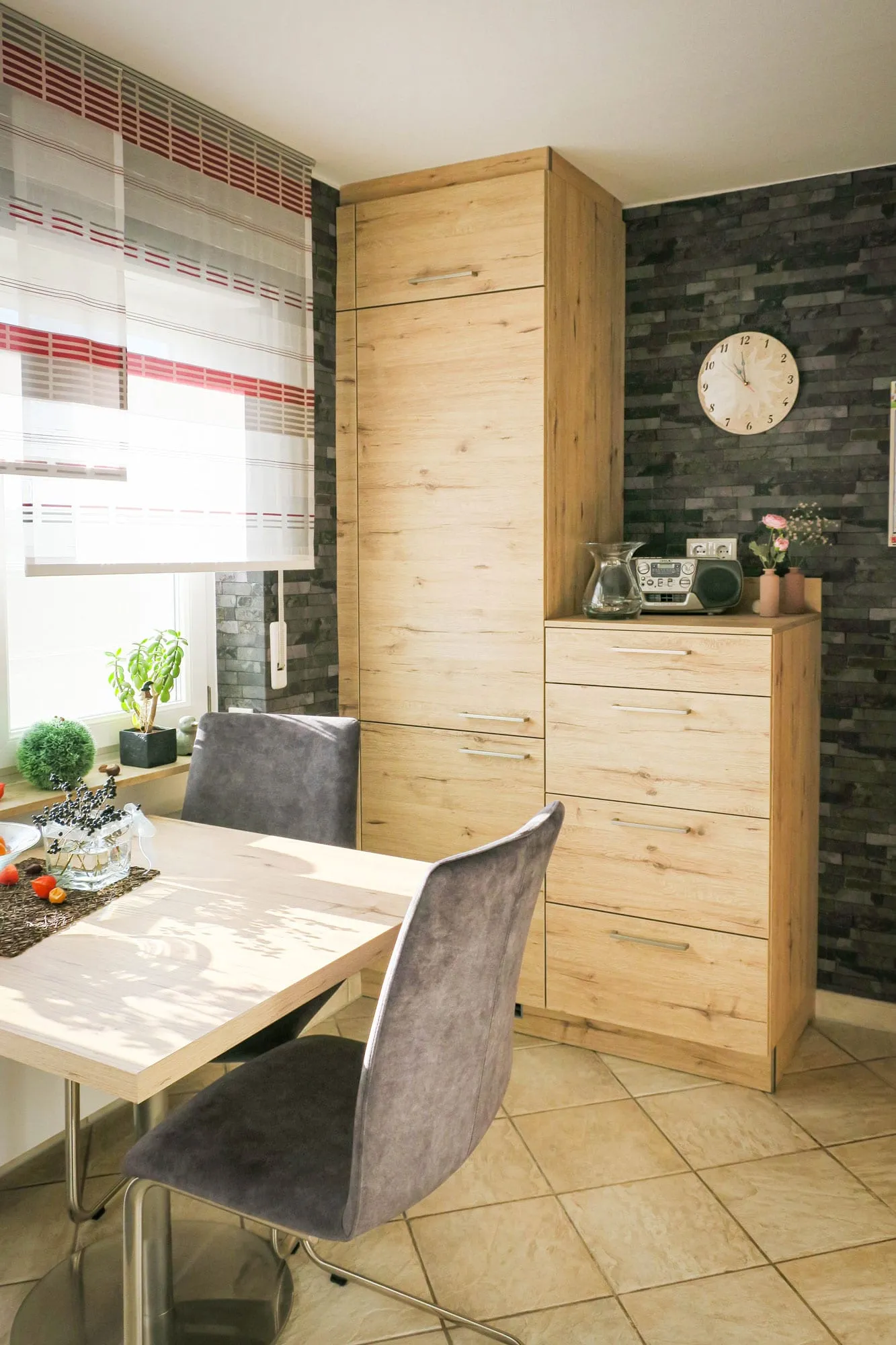 Essbereich mit Hochschrankkombination und Sideboard in Eiche-Holzoptik, grauen Esszimmerstühlen an Holztisch und Steinmosaik-Wand mit Wanduhr
