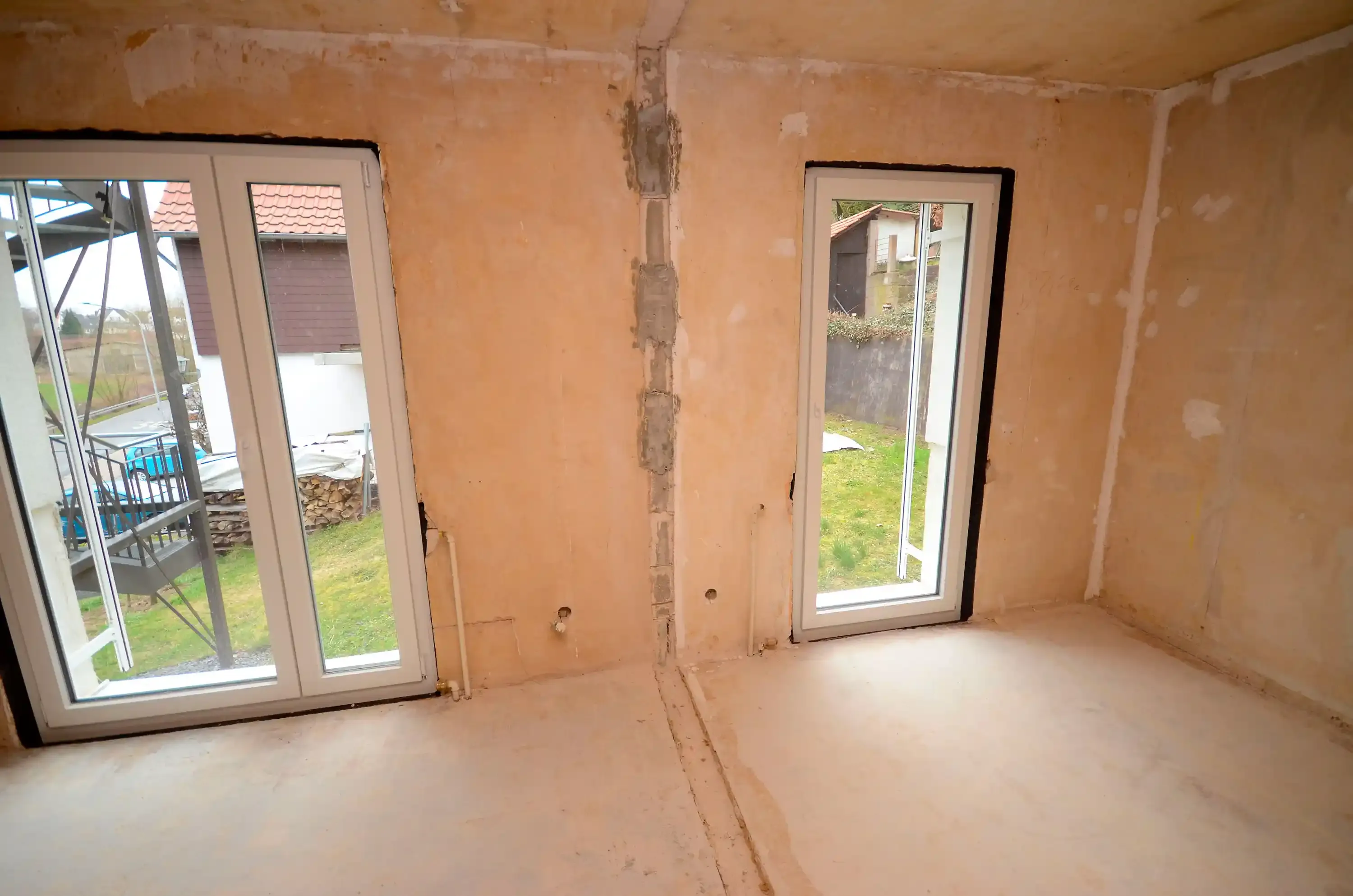 Eckraum im Rohbau mit zwei bodentiefen Fenstertüren zur Terrasse, gespachtelten Wänden, freiliegender Elektroleitung und Estrichboden – vor der Modernisierung durch Broßler Küche Aktiv (Standort Ehrbach/Großheubach)
