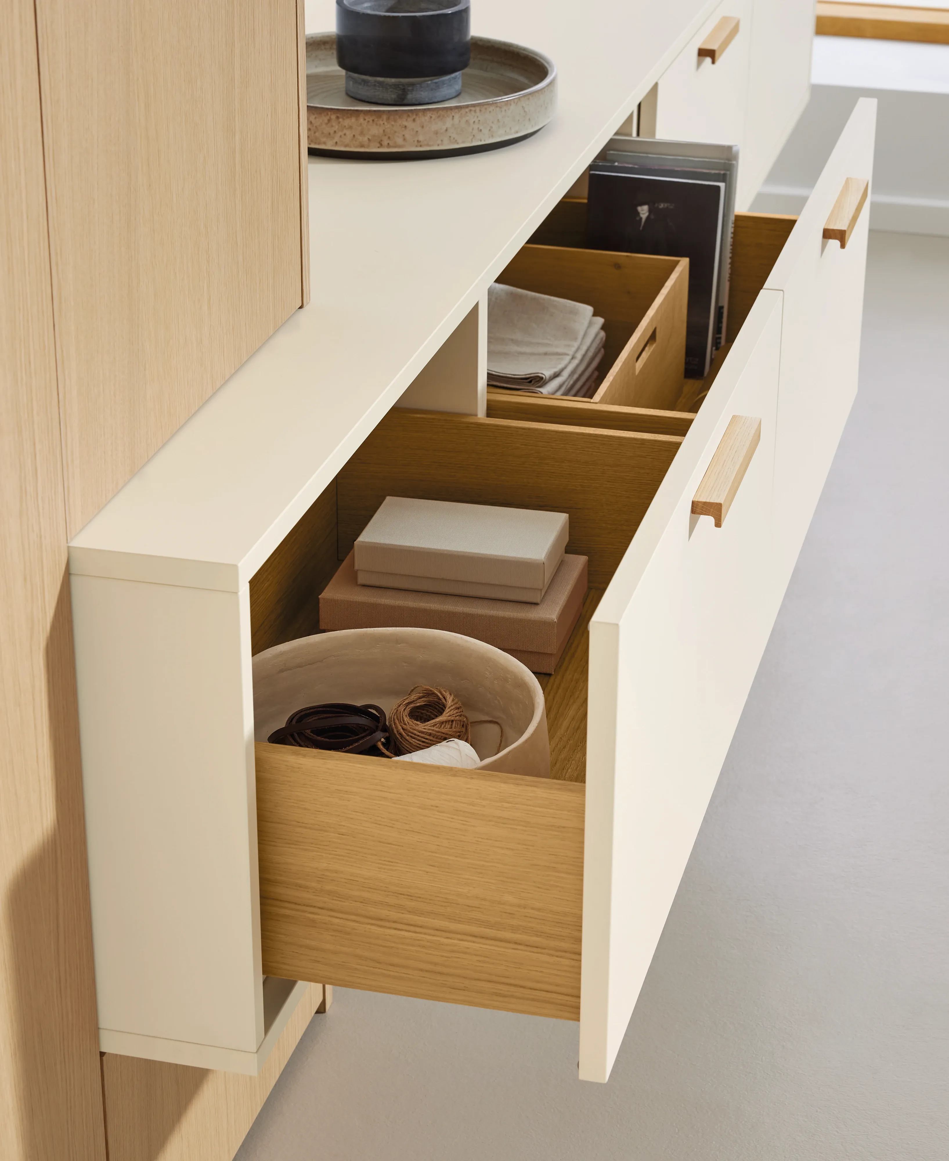Geöffnete Sideboard-Schubladen in Crème mit Holz-Griffmulden, Aufbewahrungsboxen, Schallplatten und Korb