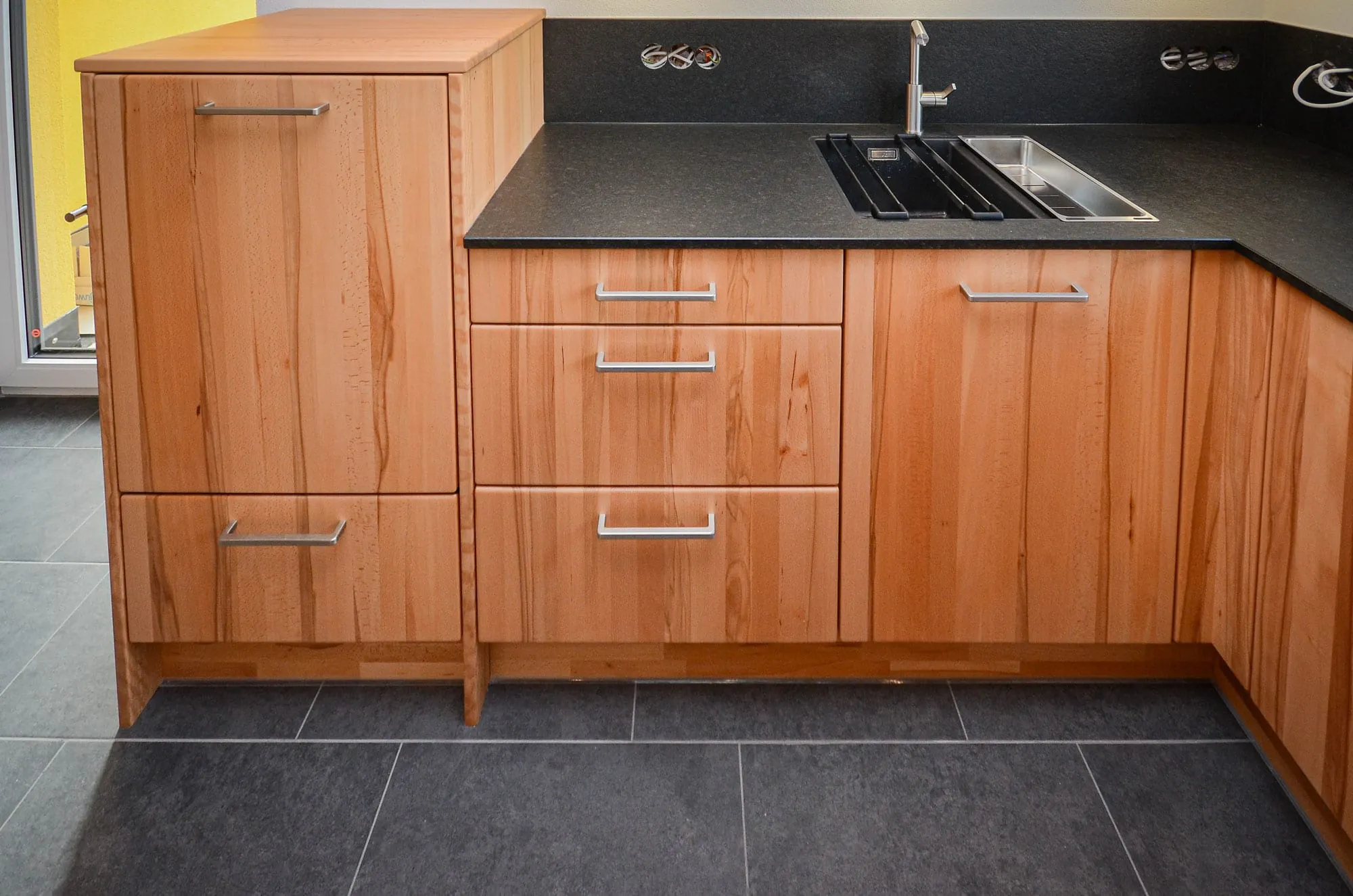 Detailaufnahme einer Massivholz-Küche mit schwarzer Steinplatte, Edelstahl-Doppelspüle und freistehenden Unterschränken auf Füßen.