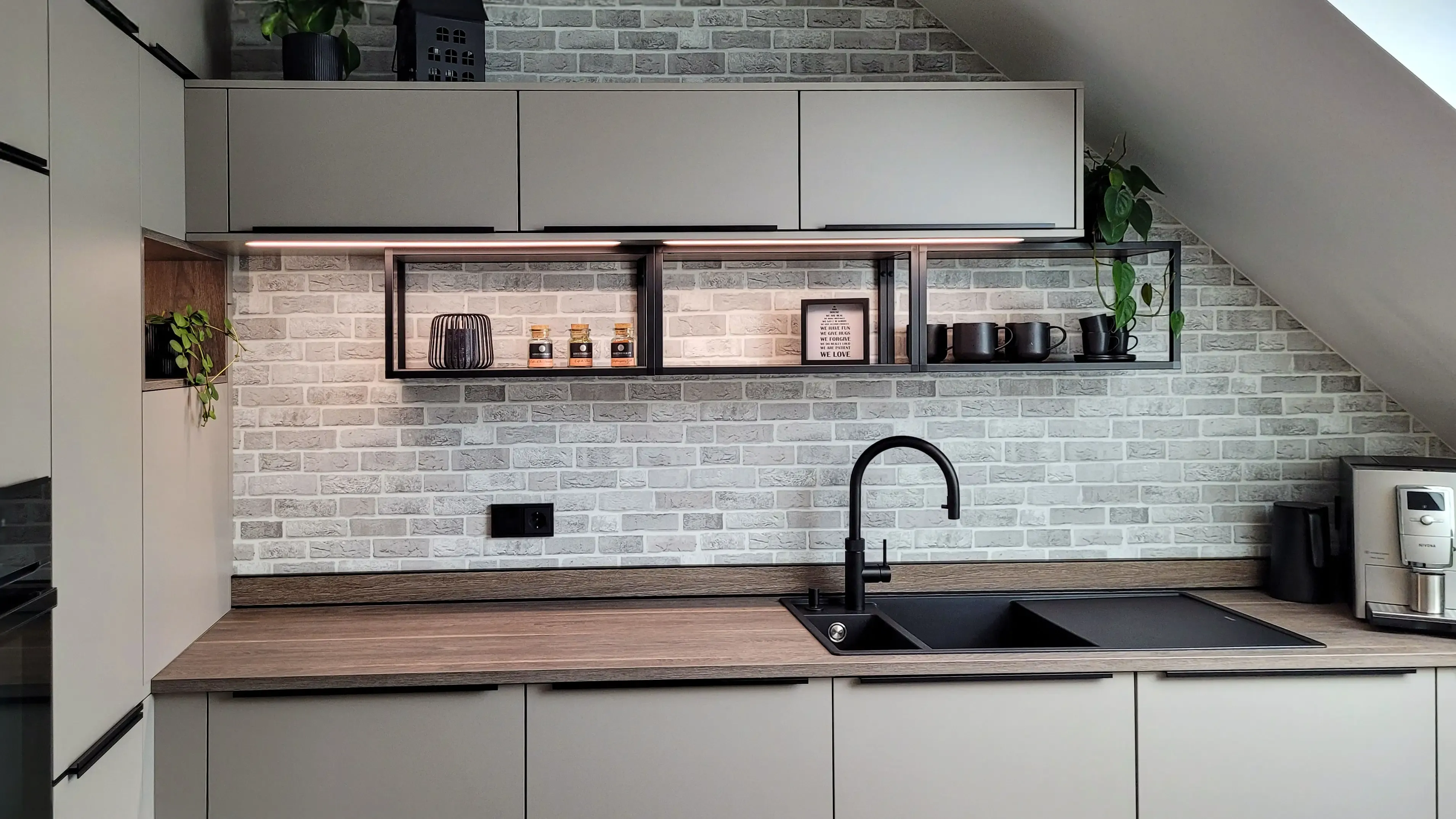 Frontale Detailansicht der Spülzone mit schwarzer Granitspüle, schwebendem Holzregal und Backsteinwand.