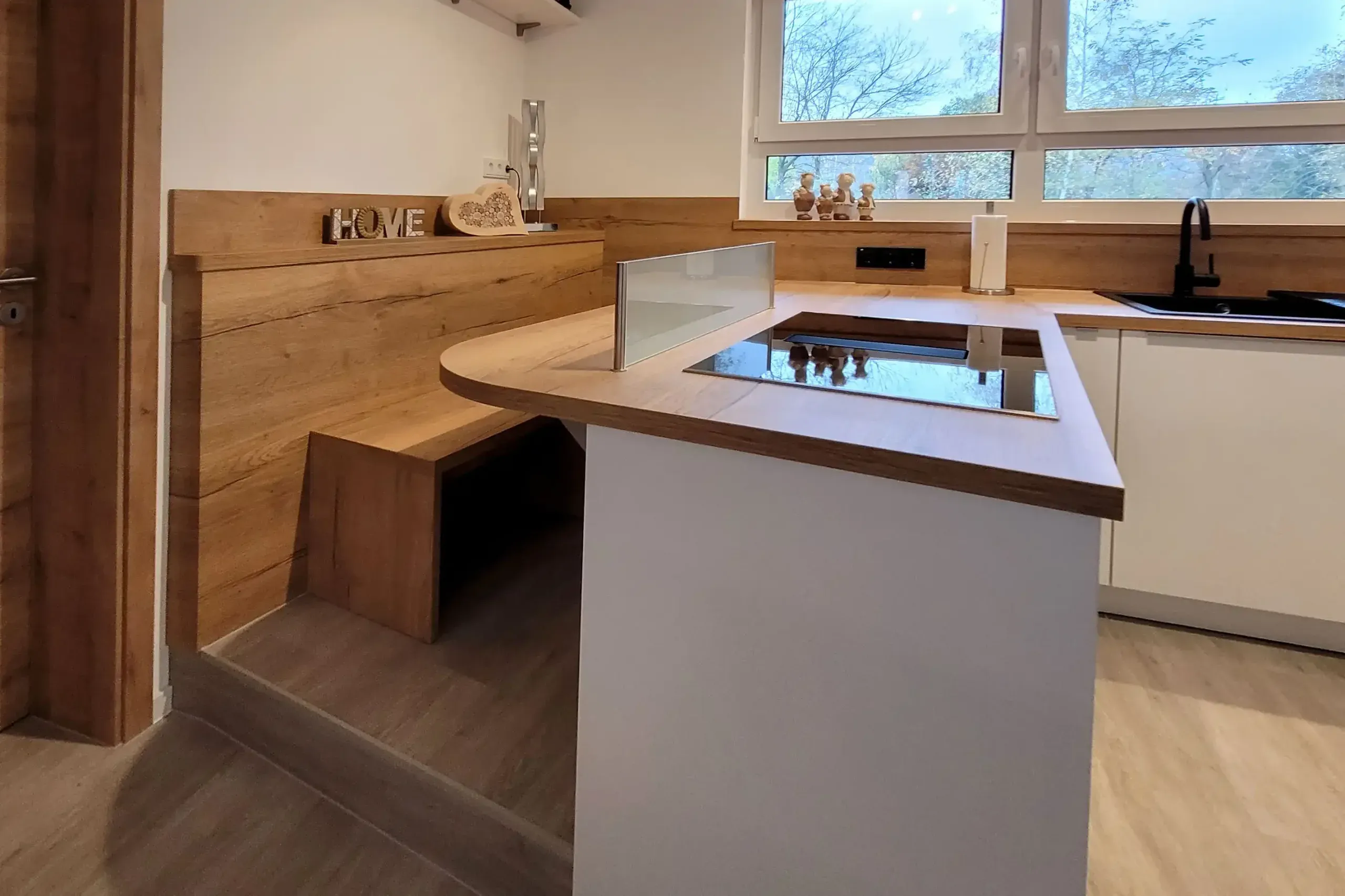 Insel mit Kochfeld und integrierter Holz-Sitzbank vor der Spüle unter dem Fenster.