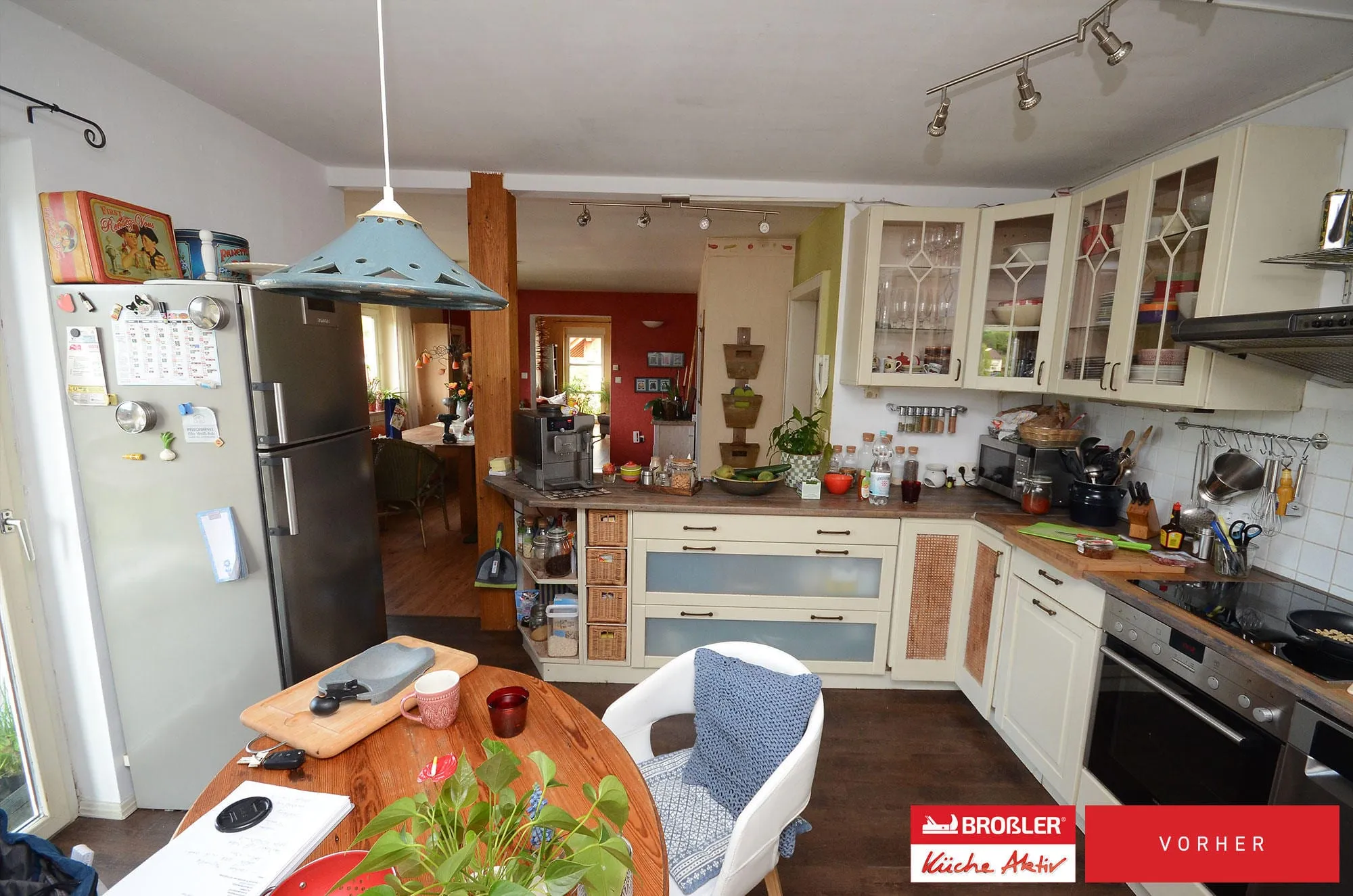 Vorher-Übersicht der Landhausküche: L-Form mit Glasvitrinen, Rattan-Einsätzen, Kühlschrank und rundem Esstisch.