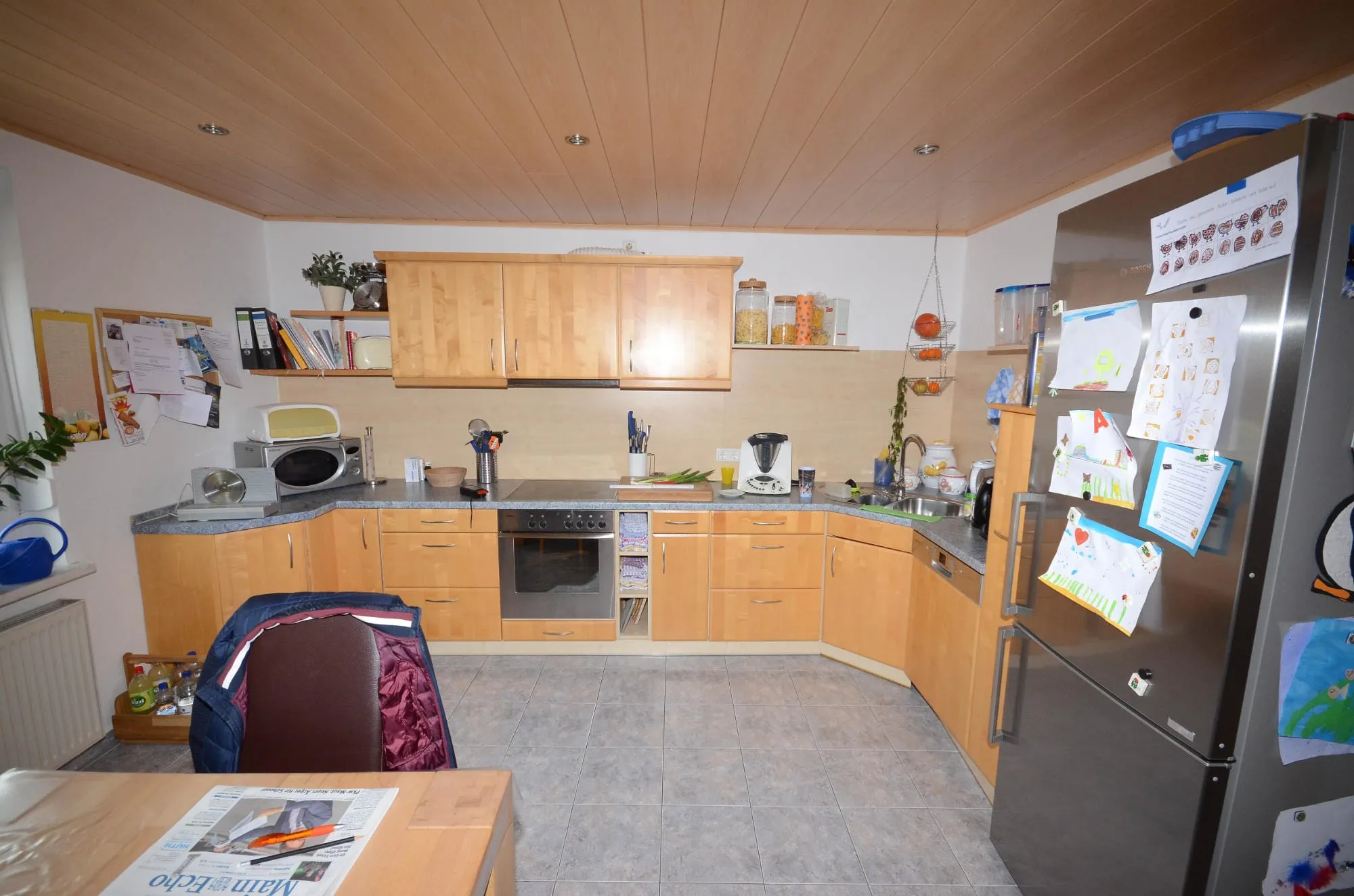 Vorher-Aufnahme einer U-Küche in heller Buche-Holzoptik mit grauer Steinplatte, Kühl-Gefrier-Kombi und Holzdecke.
