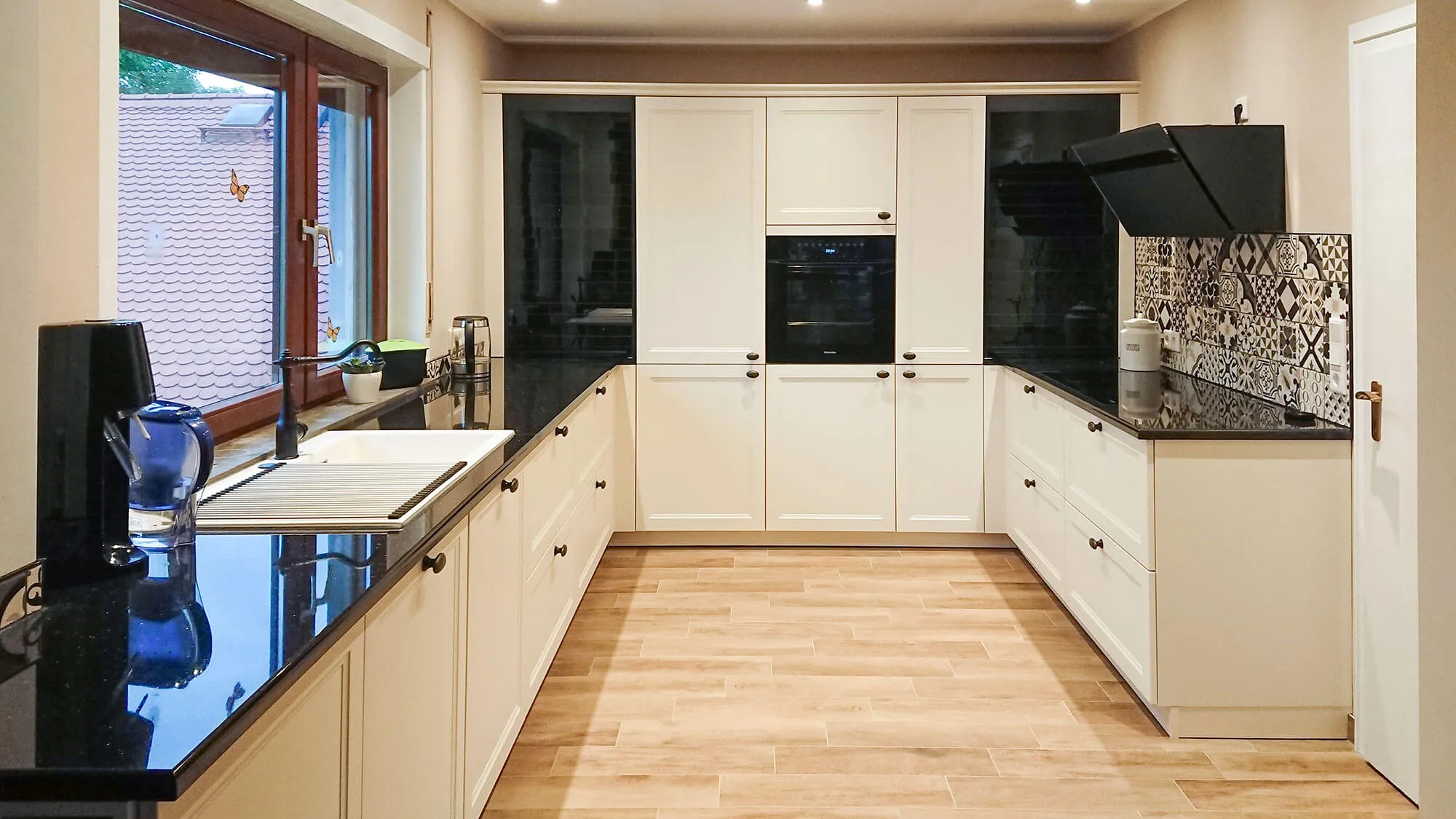 Landhaus-U-Küche mit cremefarbenen Kassettenfronten, schwarzer Arbeitsplatte und Patchwork-Spritzschutz