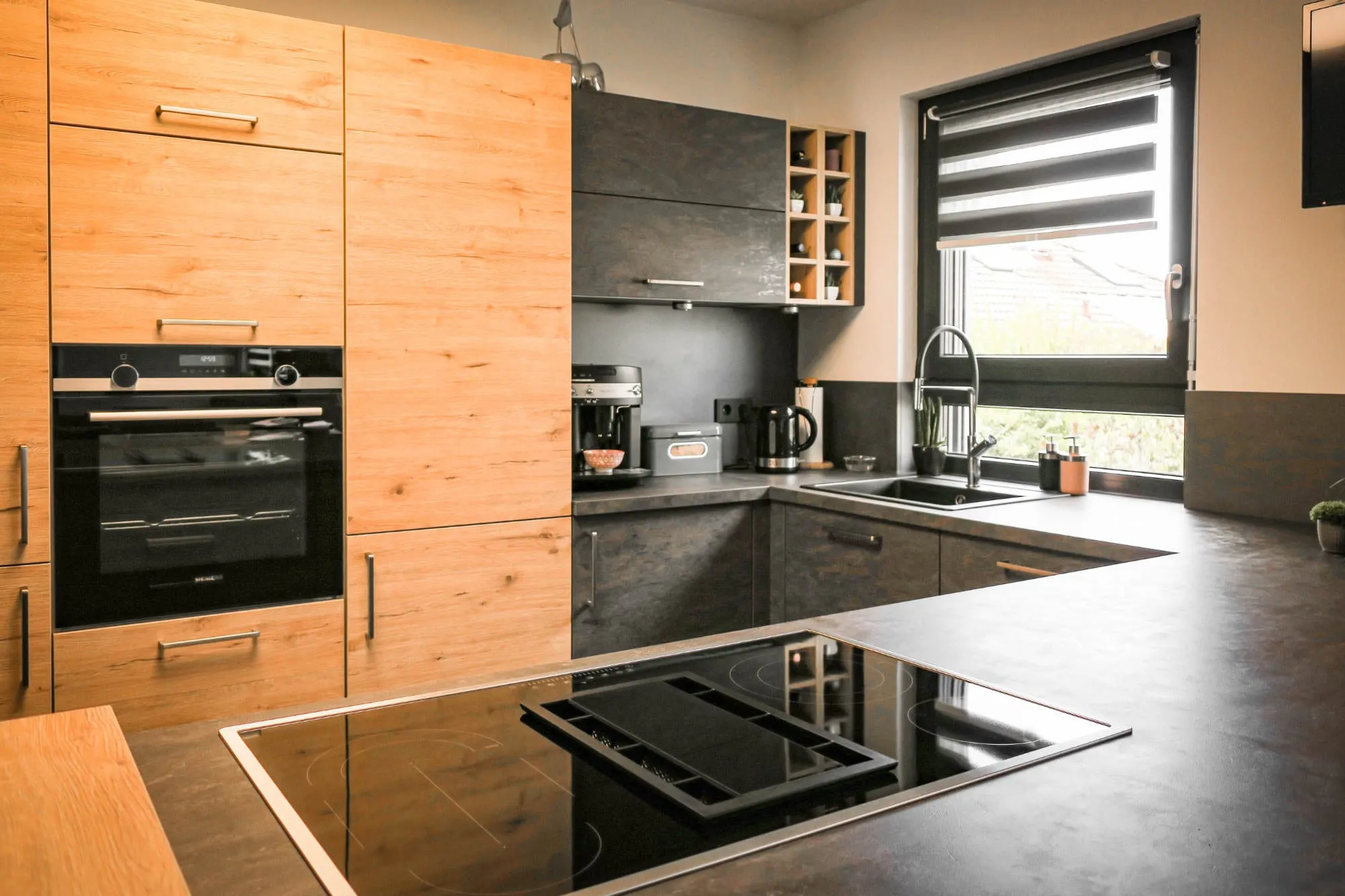 Moderne L-Küche mit Eiche-Holzfronten und anthrazitfarbenen Hochglanzfronten, integriertem Kochfeld auf der Halbinsel und schwarzer Spüle am Fenster