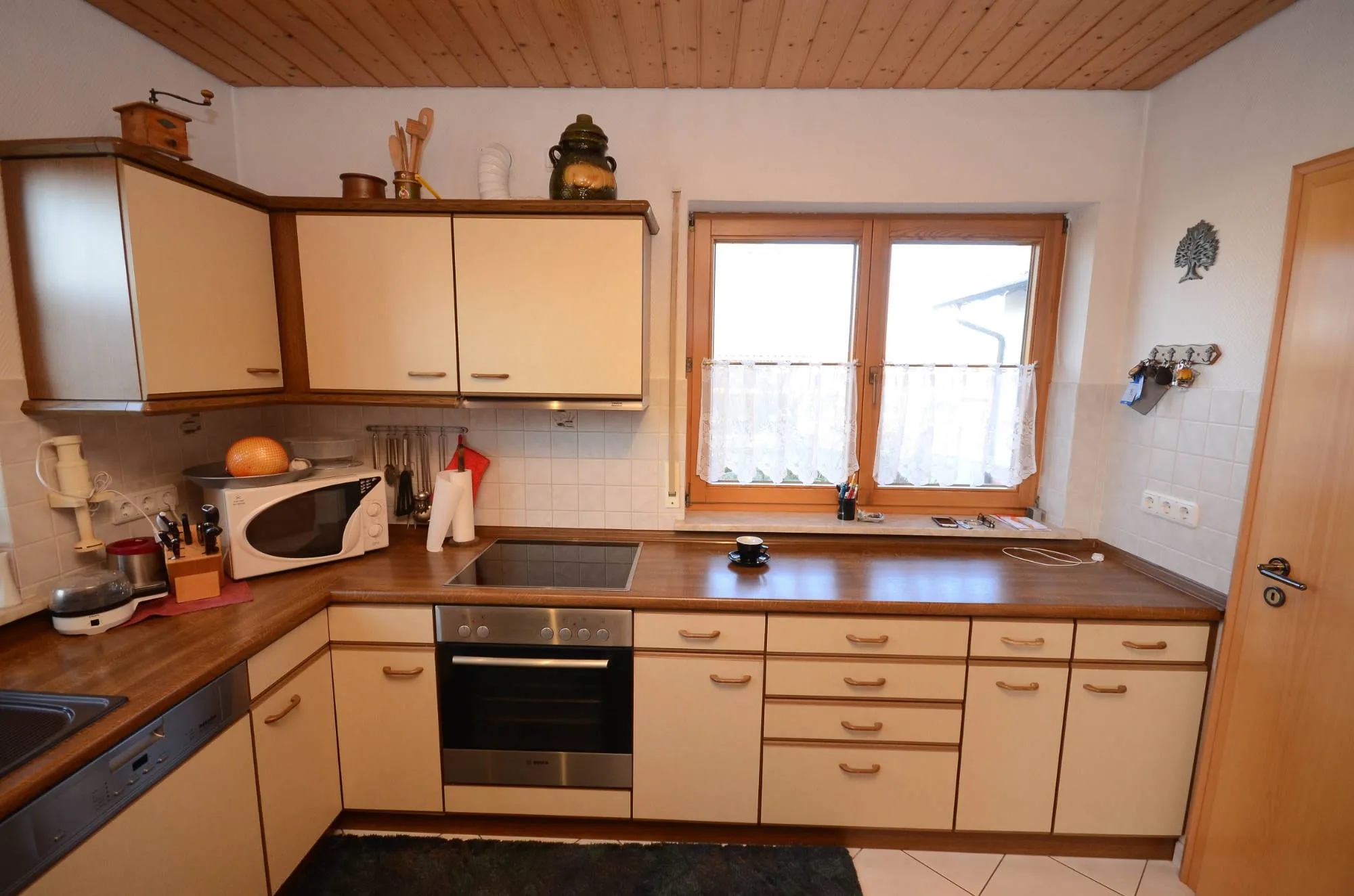 Vorher-Aufnahme der alten Küche: Arbeitszeile mit Einbau-Backofen, Kochfeld und Mikrowelle vor Holzdecke und Fliesenboden.