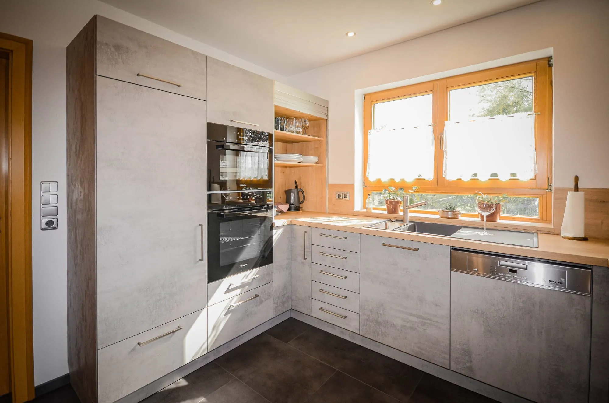 Eckperspektive der U-Küche in Beton-Steinoptik mit Hochschrank-Backofen, Spüle unter Fenster und offenem Holz-Eckregal.