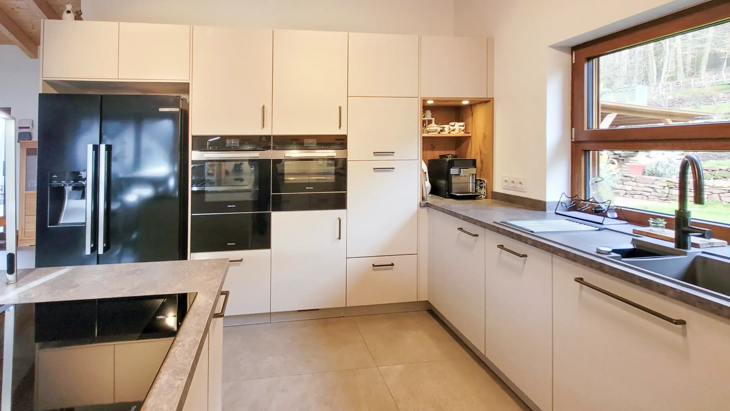 Grifflose Hellbeige-Küche mit Side-by-Side-Kühlschrank, Hochschrankzeile und Fenster-Spüle.