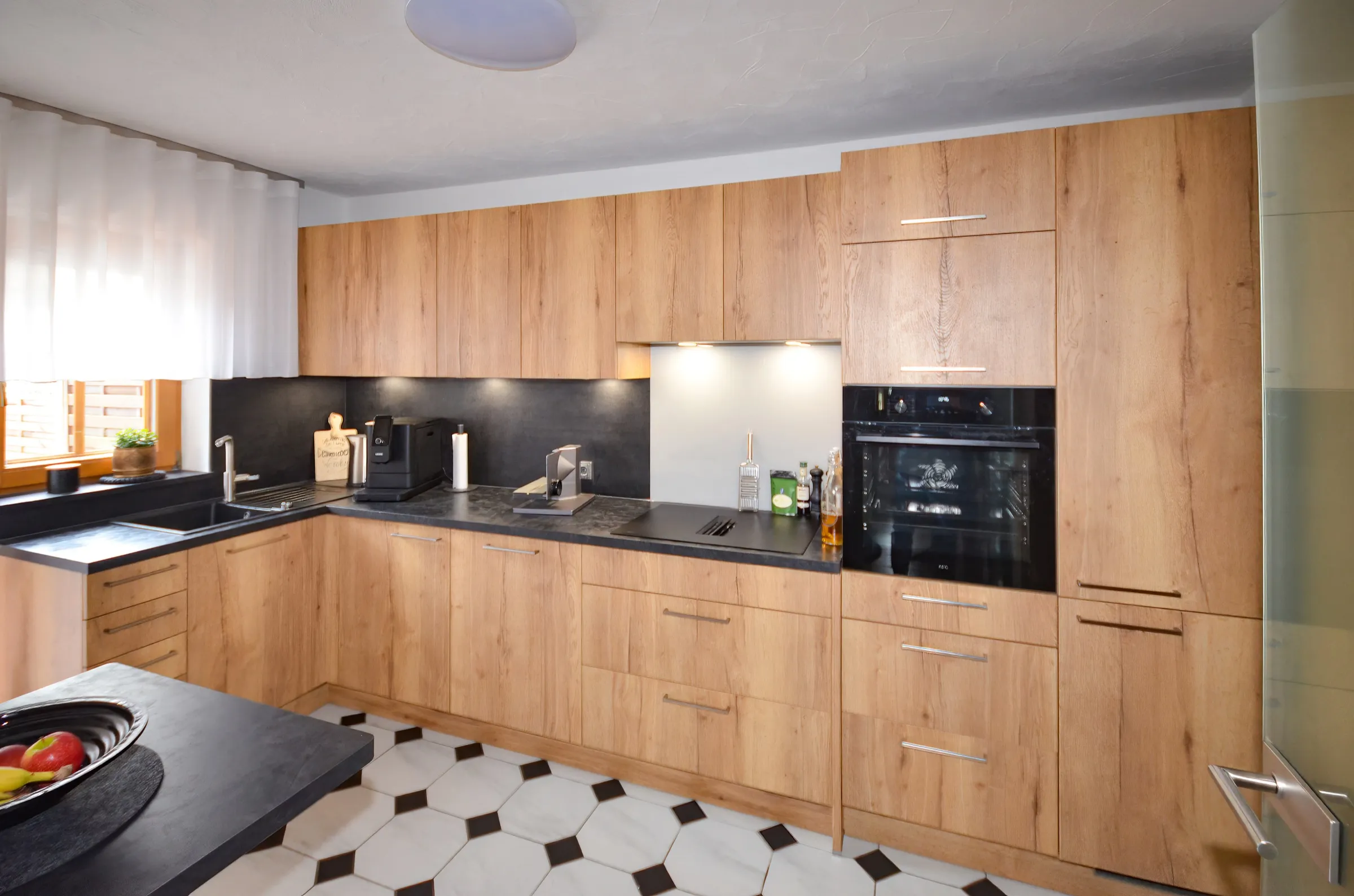 Renovierte Küche mit hellen Holzfronten, dunkler Arbeitsplatte, dunkler Nischenrückwand und hochgebautem Backofen
