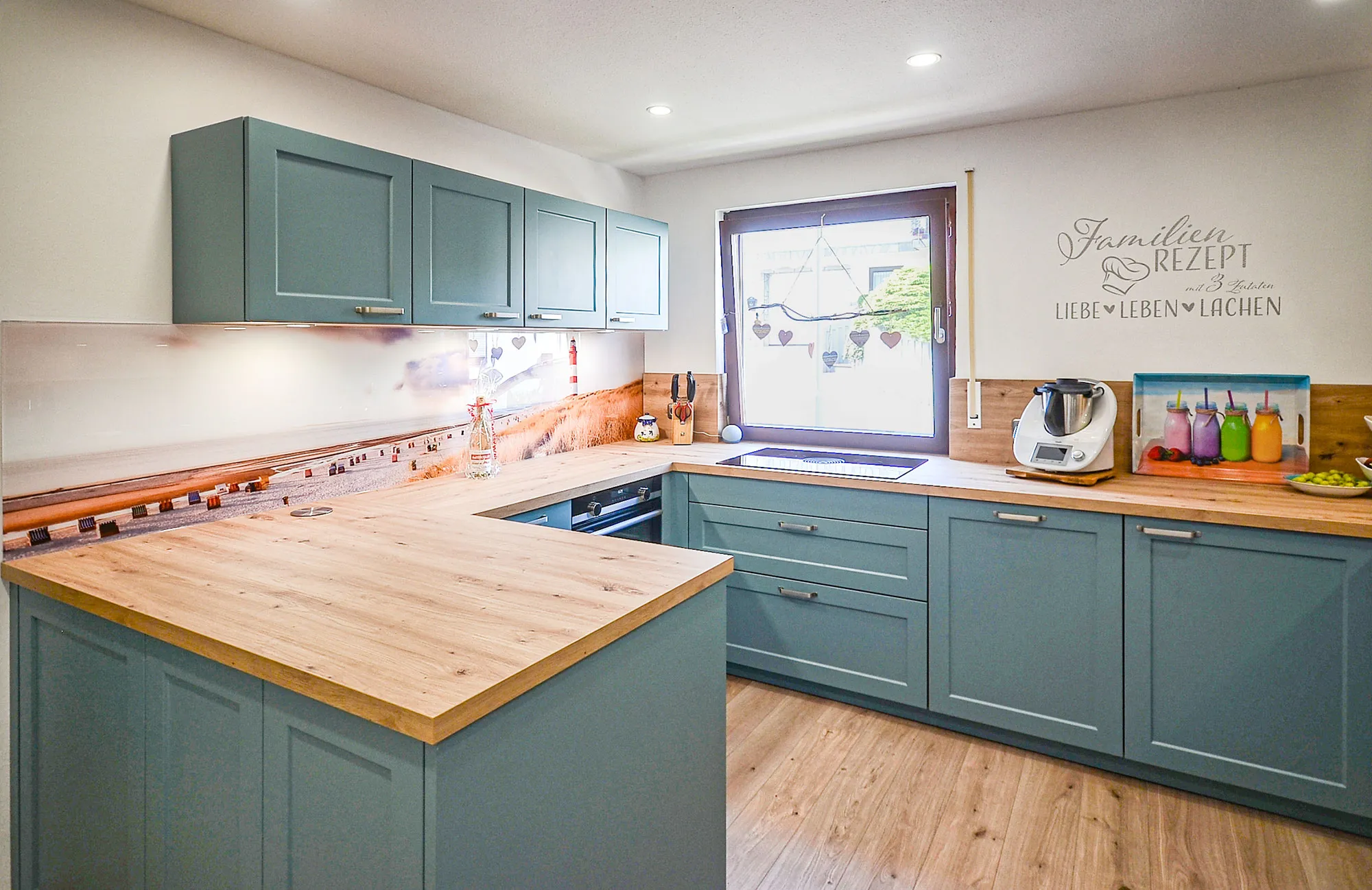 U-Küche im Landhausstil mit hellblauen Kassettenfronten und massiver Arbeitsplatte in warmer Holzoptik, Nischenrückwand mit Strand-Motiv, Glaskeramik-Kochfeld unter dem Fenster, Wandtattoo 'Familien-Rezept' – Küchenplanung von Dietmar Kraus für Broßler Küche Aktiv