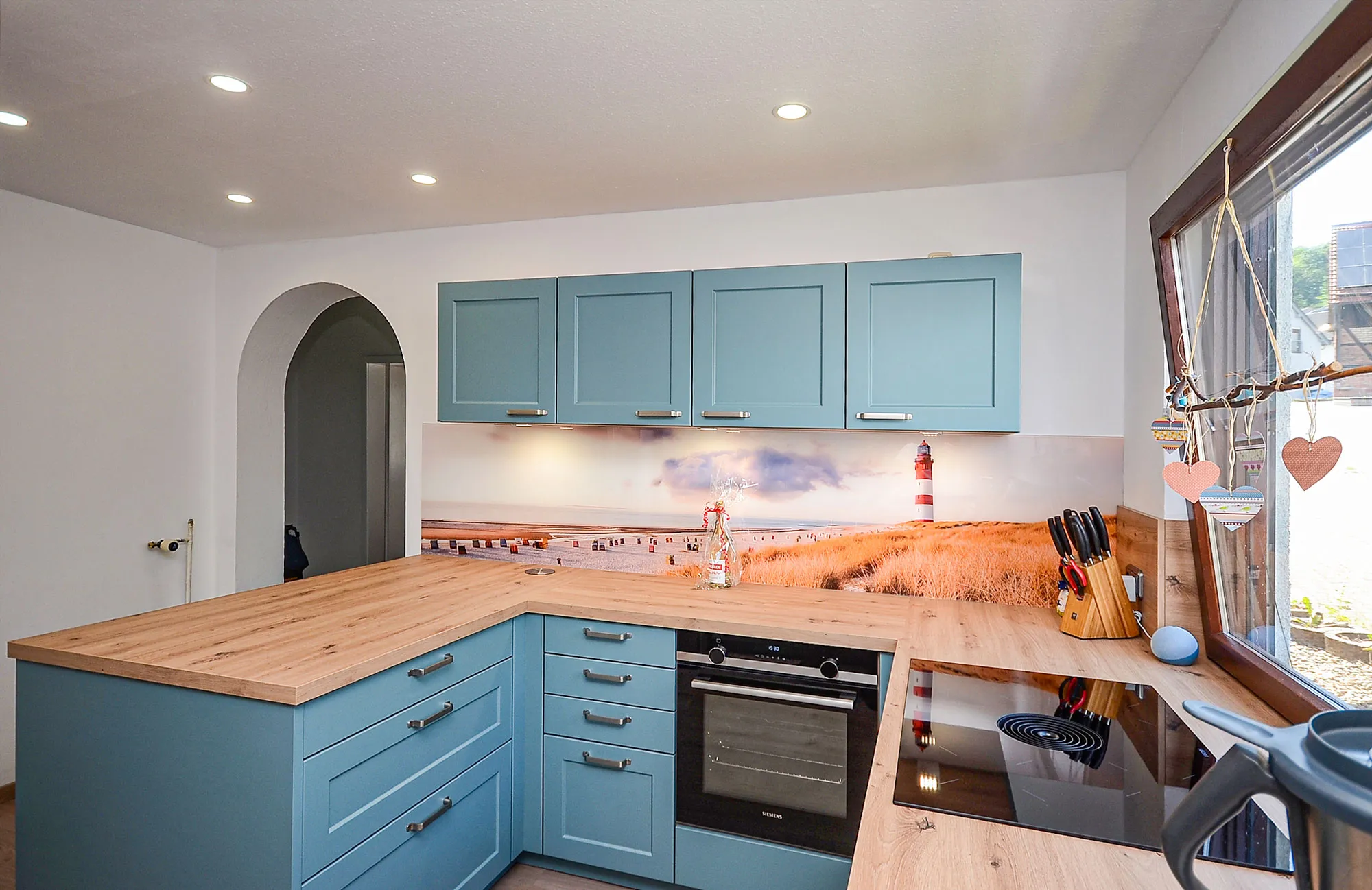 L-Küche im Landhausstil mit hellblauen Kassettenfronten und massiver Arbeitsplatte in warmer Holzoptik, integrierter Backofen und Glaskeramik-Kochfeld, Nischenrückwand mit Strand- und Leuchtturm-Motiv, Rundbogen-Durchgang links und Fenster mit herzförmiger Deko rechts – Küchenplanung von Dietmar Kraus für Broßler Küche Aktiv